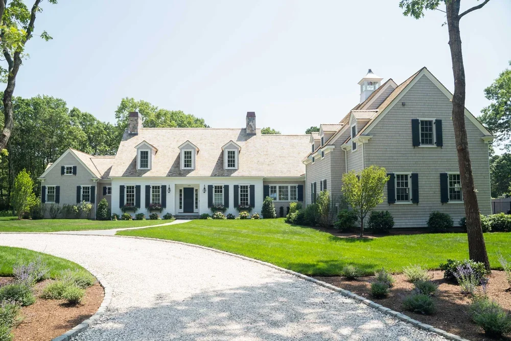 Front exterior of shingle-style family compound in Oyster Harbors, Osterville, featuring a central main house with dormers and black shutters, attached carriage house wing with cupola, twin stone chimneys, and gravel drive through landscaped grounds.