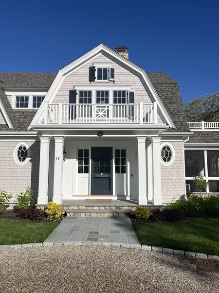 Front entrance of shingle-style coastal home featuring a gambrel roof gable, second-floor balcony, columned portico, blue front door, oval windows on flanking wings, and bluestone walkway through gravel drive. James Phillip Golden Architect