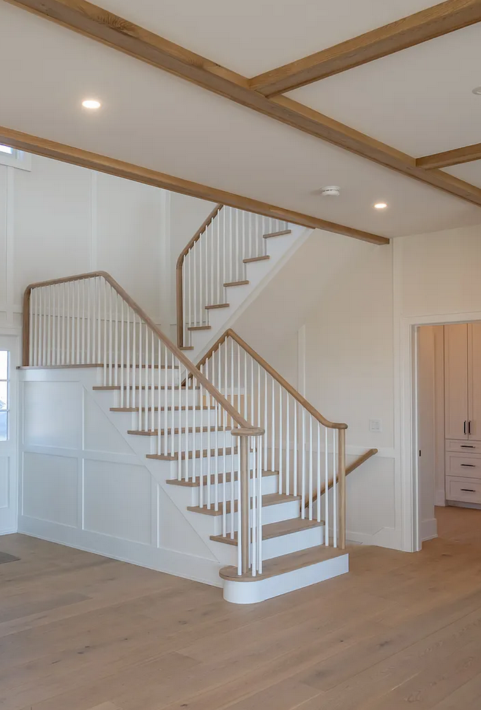 Central staircase with white-oak treads and handrail, painted spindles, board-and-batten wainscoting, and a coffered wood-beam ceiling above.