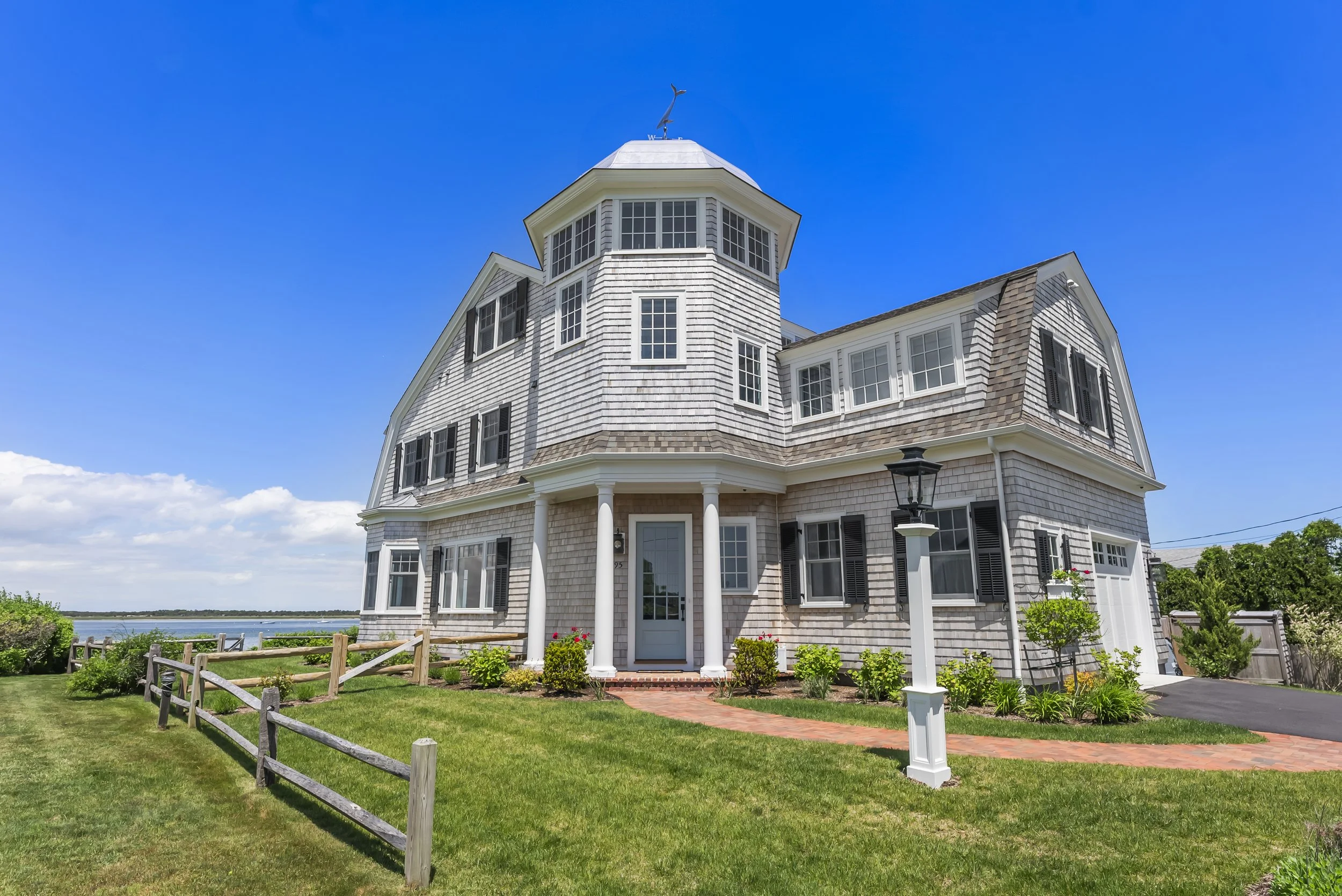 Front exterior of new construction shingle-style home on Sunset Lane, Barnstable, featuring an octagonal cupola tower topped with a whale weathervane, gambrel roof, black shutters, columned entry portico, and bay views. James Phillip Golden Architect