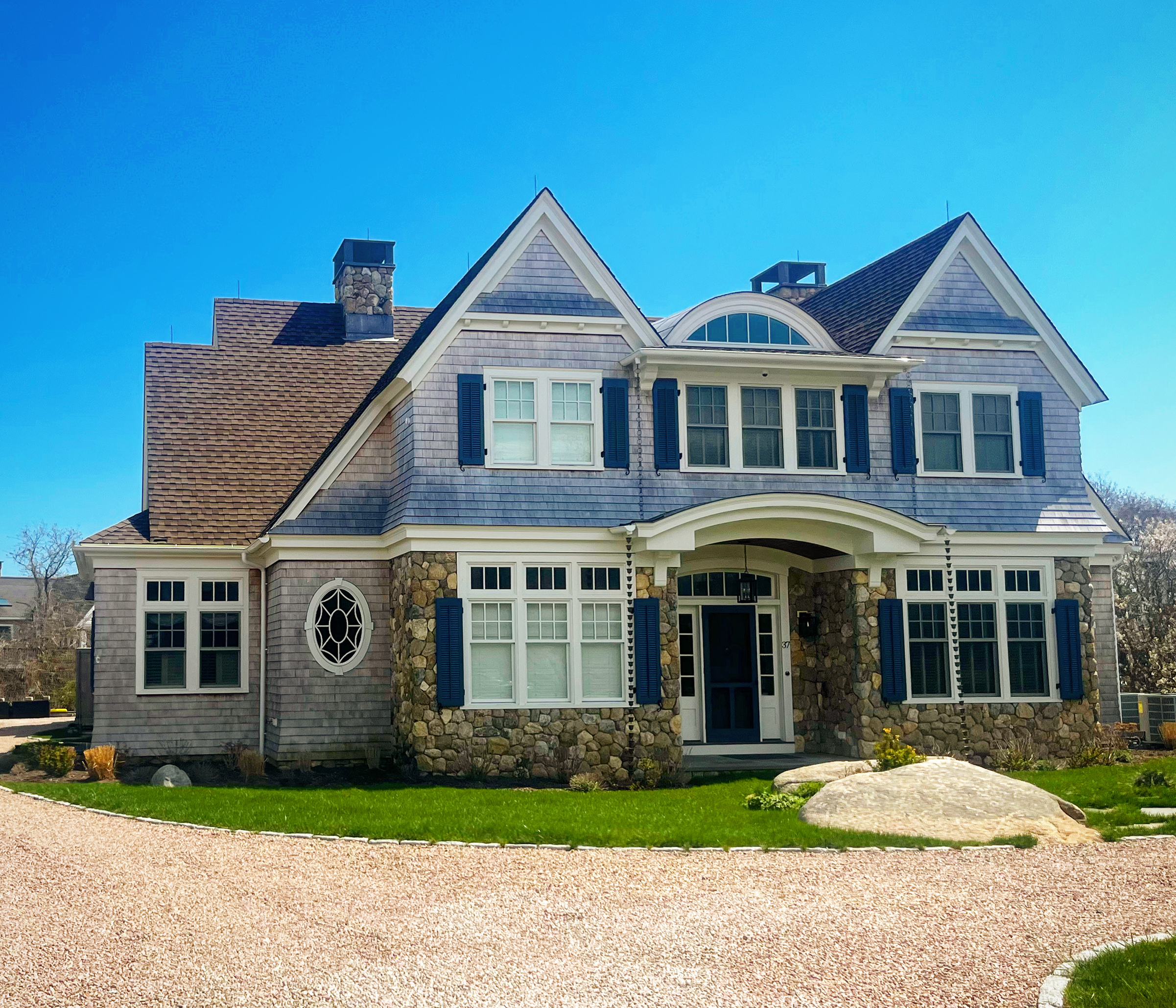 Front exterior of shingle-style home on Neptune Avenue, Charlestown, Rhode Island, featuring a fieldstone foundation and entry surround, twin front gables with eyebrow dormer between, blue shutters, decorative oval window with tracery, and crushed st