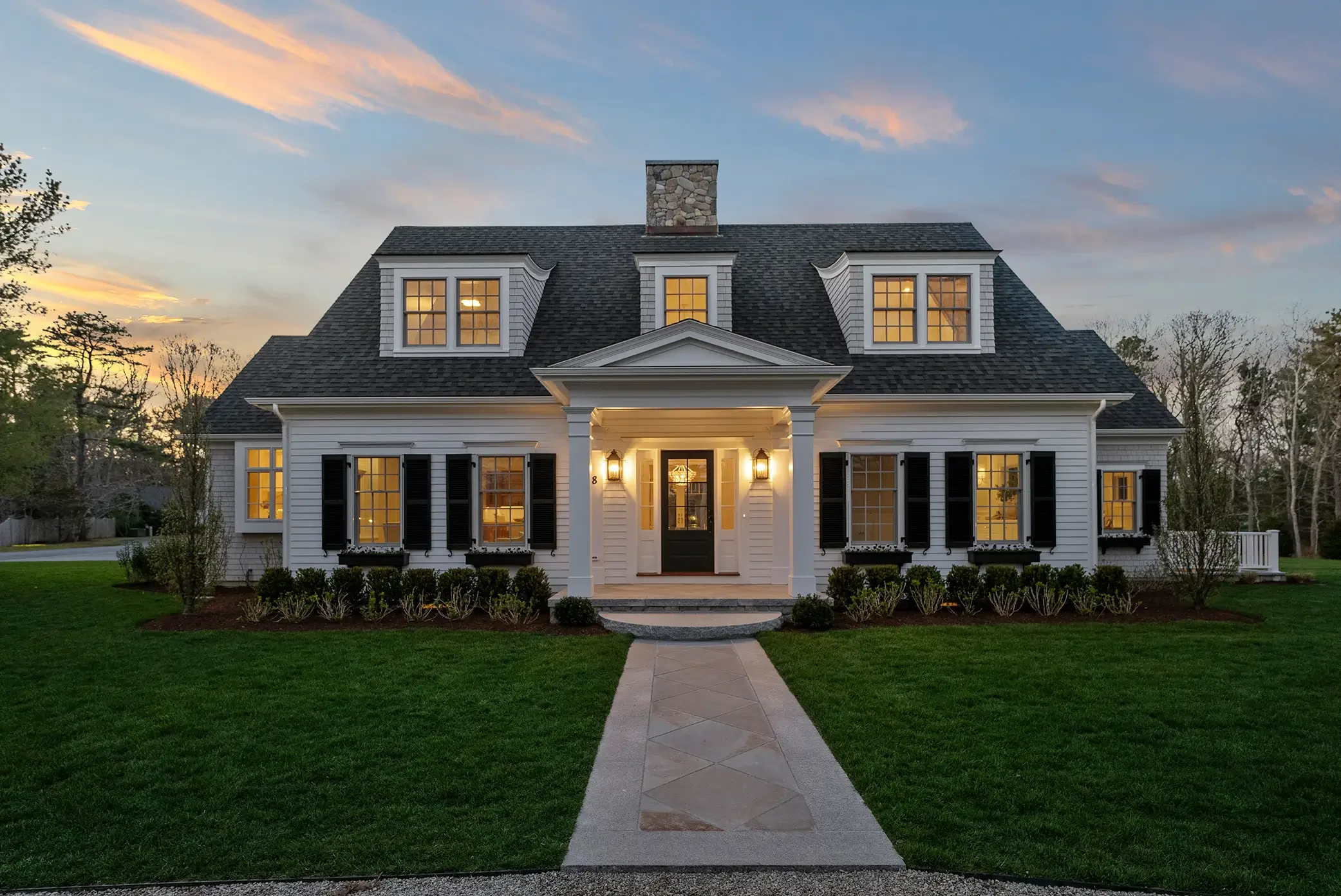 Cape Cod shingle-style home at twilight, warm interior lights glowing through windows, centered front portico with columns, gambrel roof with three dormers, stone chimney, and symmetrical landscaped front lawn. James Phillip Golden Architect