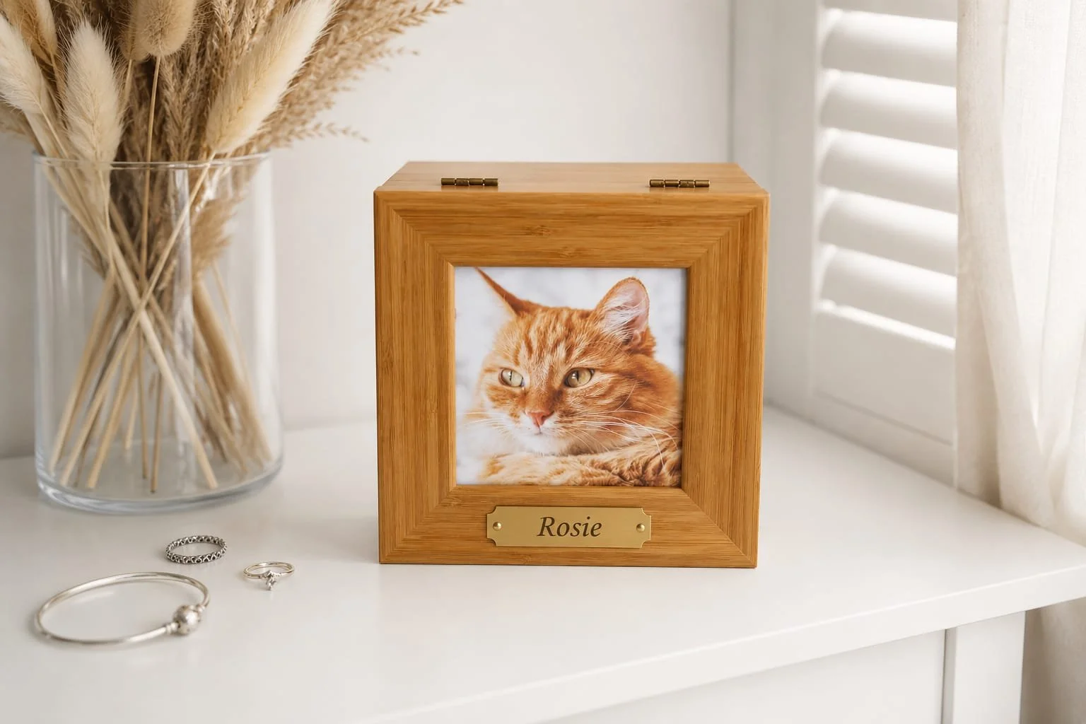 A framed photo of an orange tabby cat labeled Rosie, placed on a white surface next to a glass vase with dried pampas grass and three pieces of jewelry.