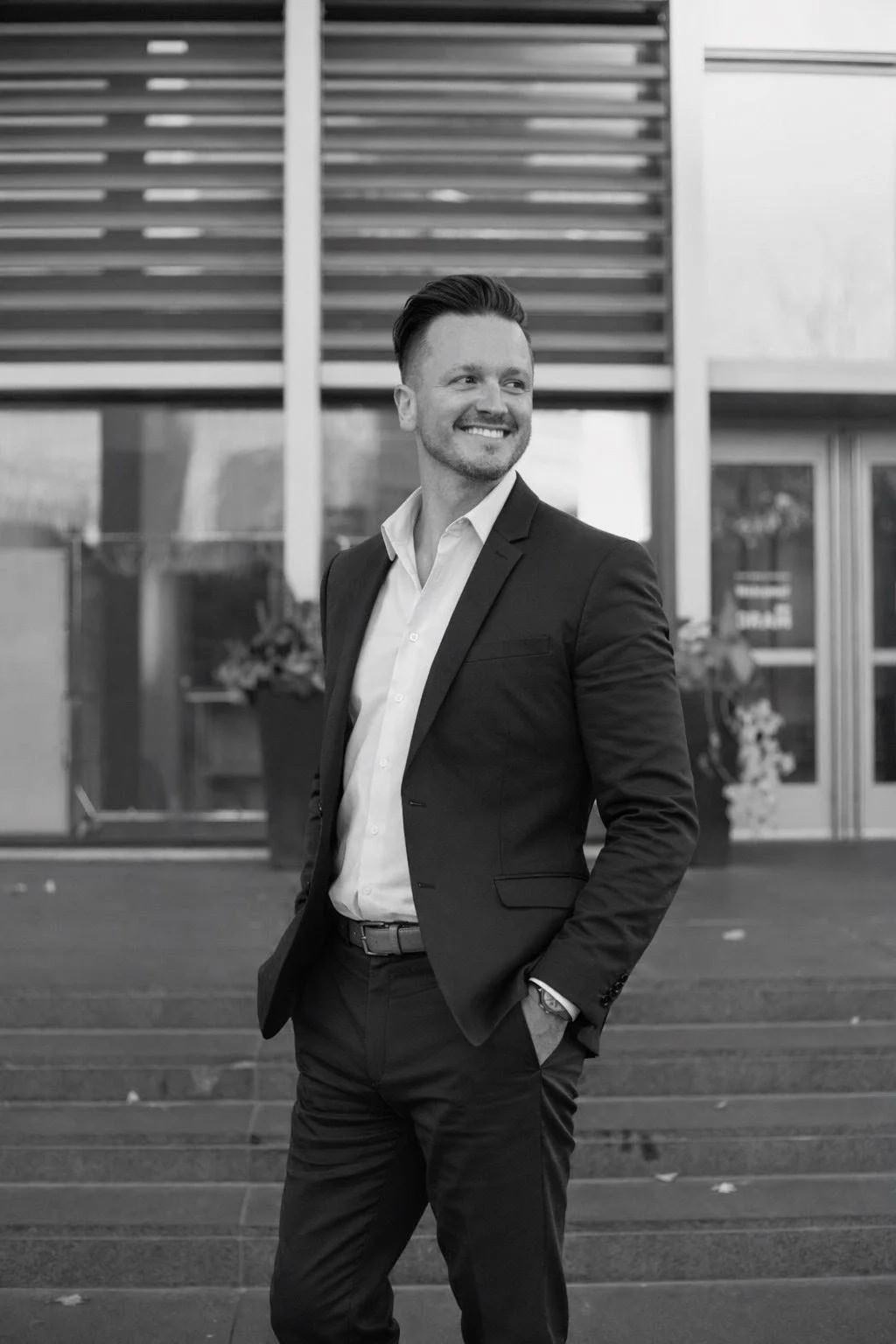 A man in a suit smiling outdoors in front of a modern building.