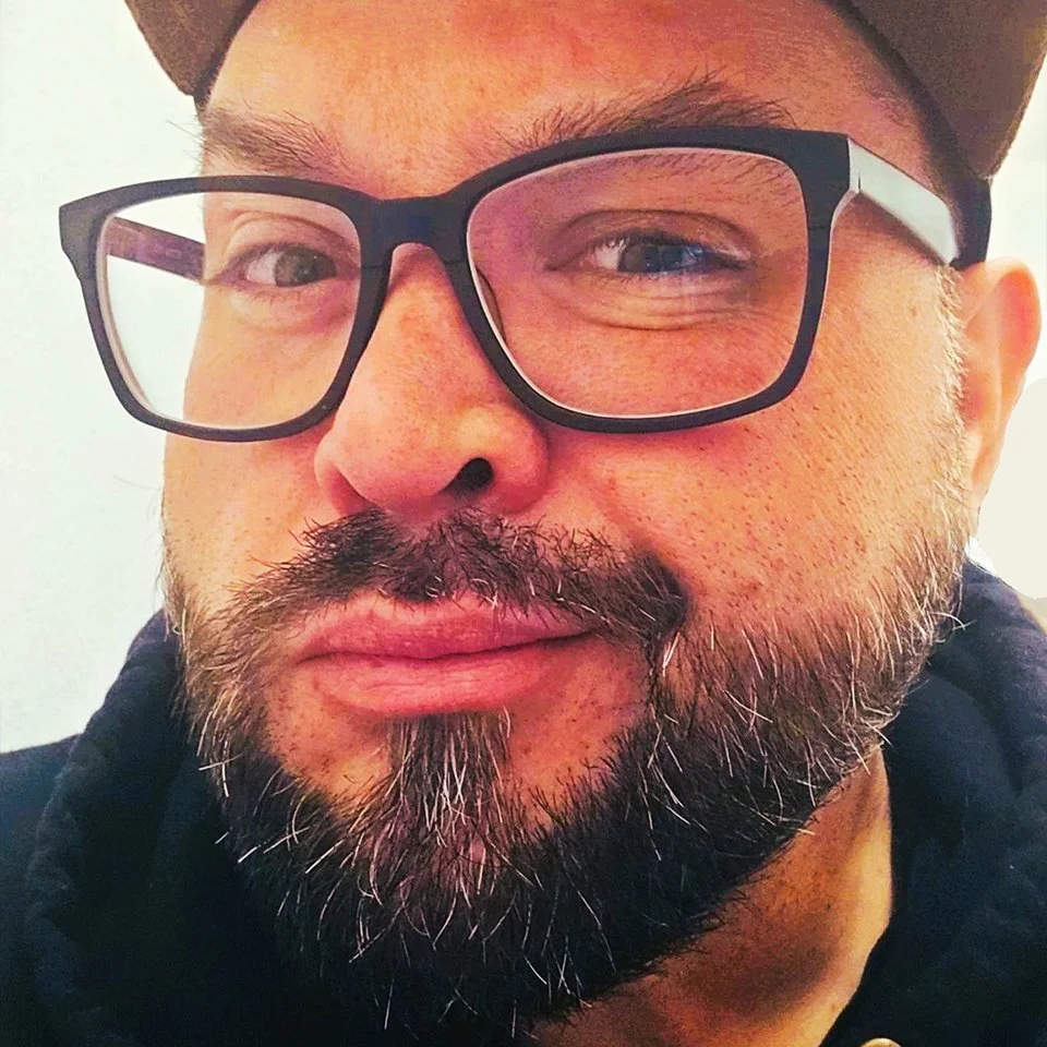 Close-up of a man with glasses and a beard, smiling slightly, wearing a cap.