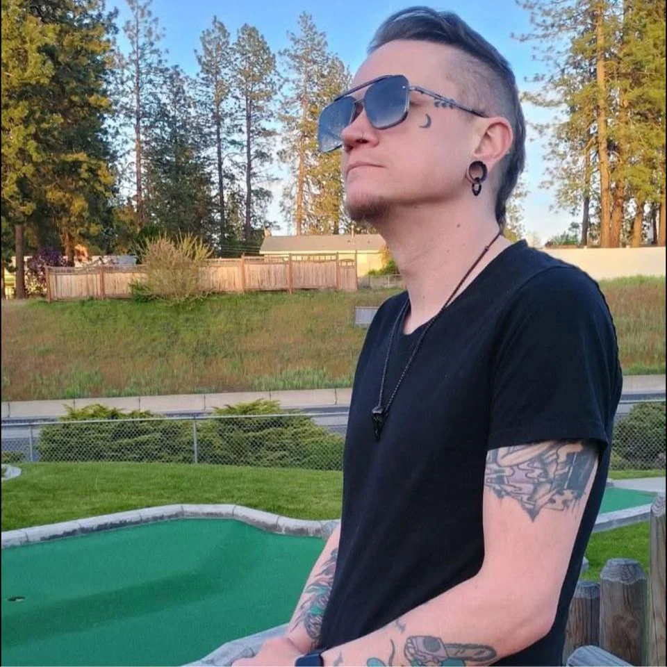 A man with dark sunglasses, a lip ring, and tattoos on his arms is standing outside near a mini golf course, with a grassy area and trees in the background.