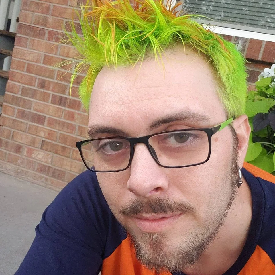 Portrait of a young man with bright green and yellow spiked hair, black glasses, and a goatee, wearing a blue and orange shirt, outdoors in front of a brick wall and greenery.