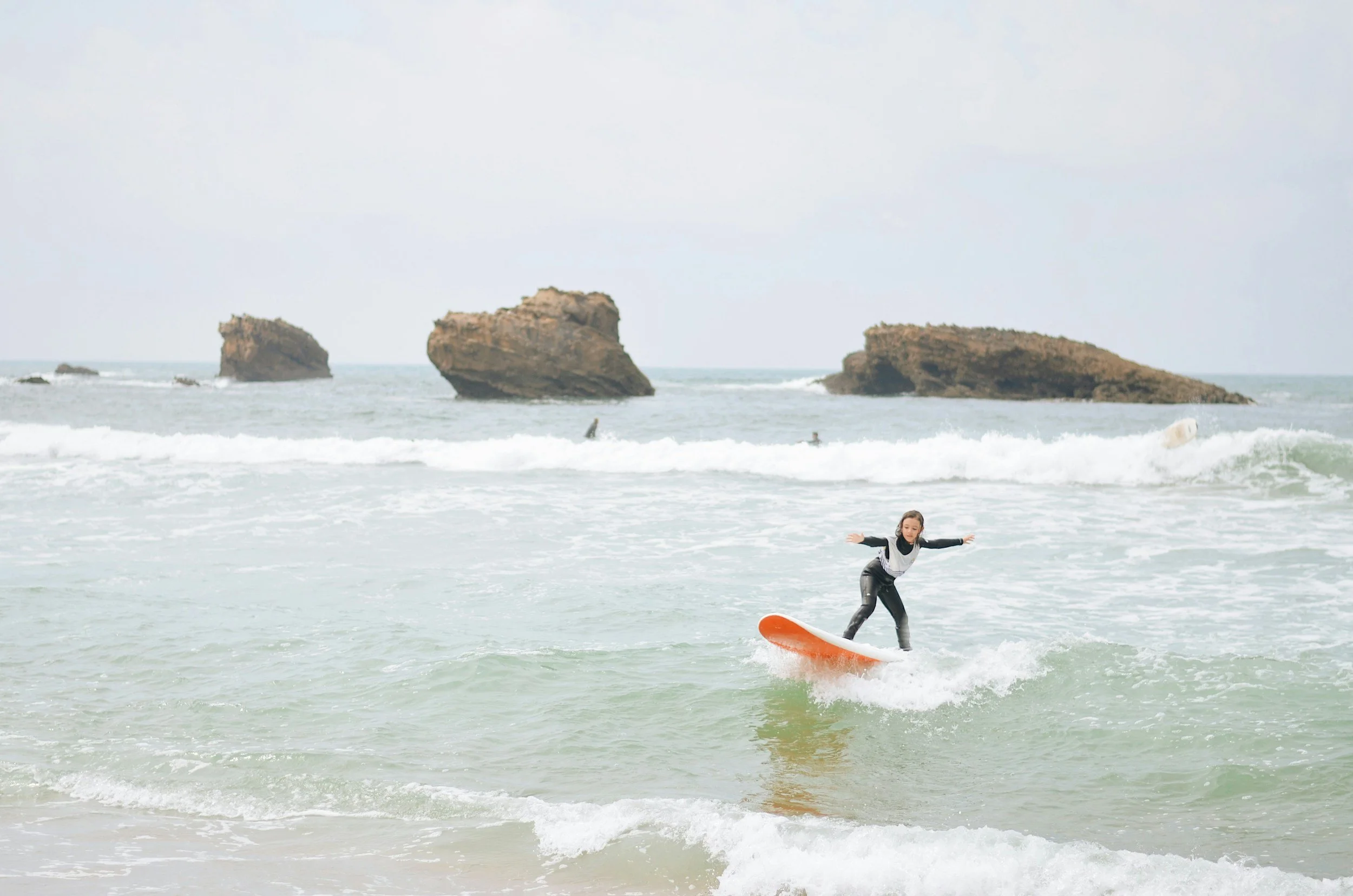 Child surfing on an orange surfboard at the beach with rock formations in the background.