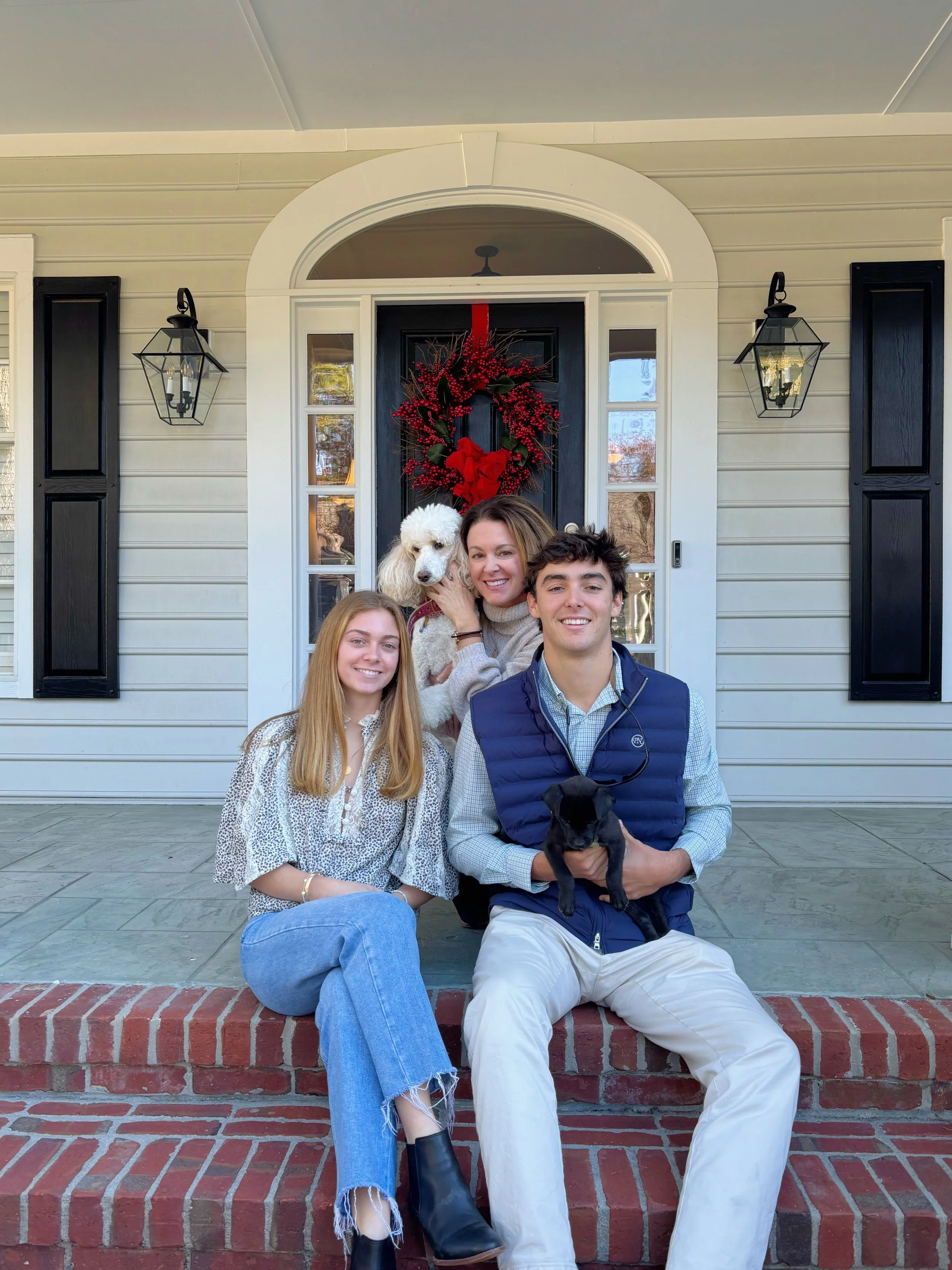 A family and their dog sitting on the steps of a house with a decorated front door for the holidays.