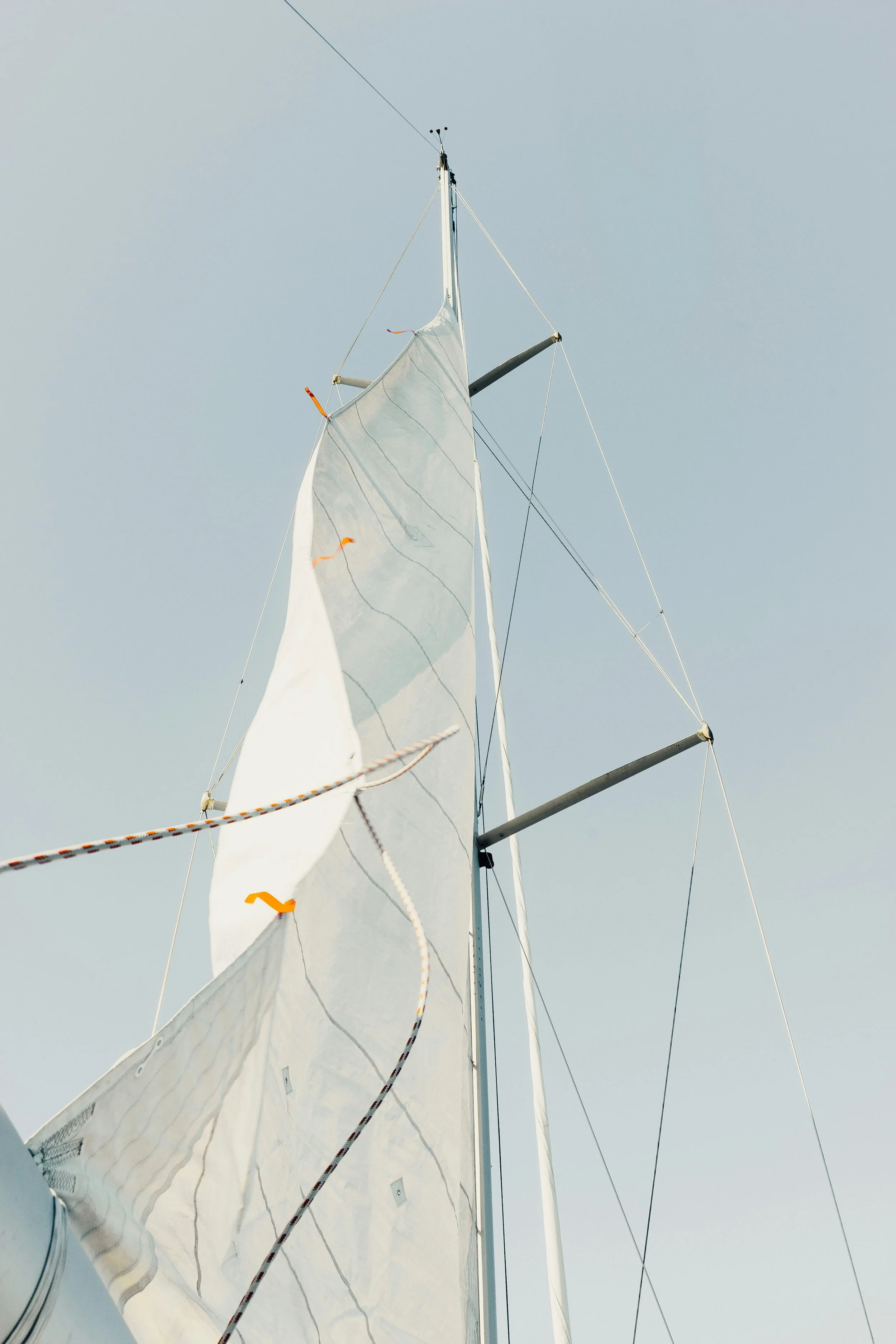 Upward view of a sailboat's mast, sail, and rigging against a clear sky.