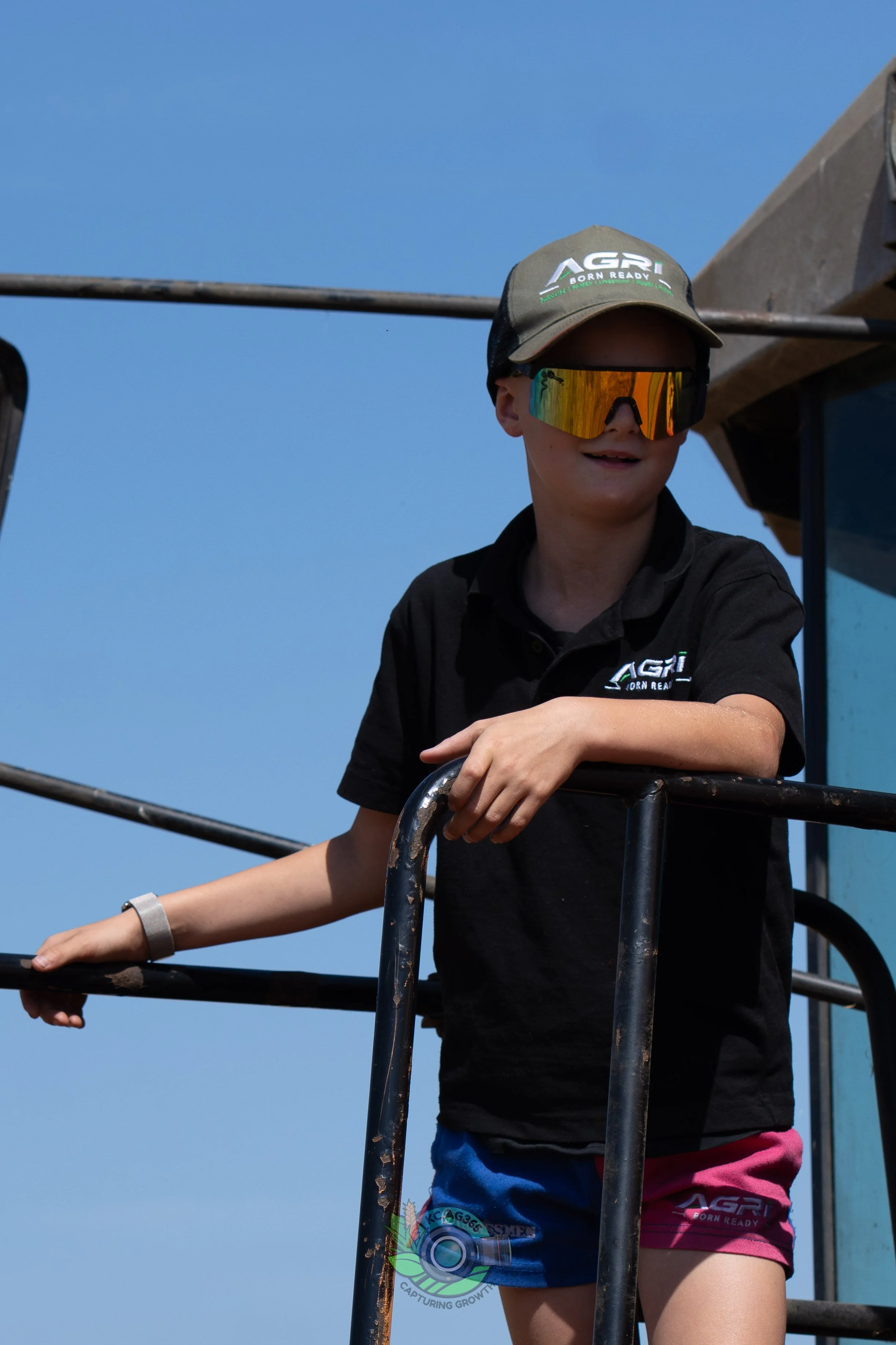 Young person wearing a gray cap with the words "AGR Born Ready" and reflective sunglasses, standing on a combine harvester