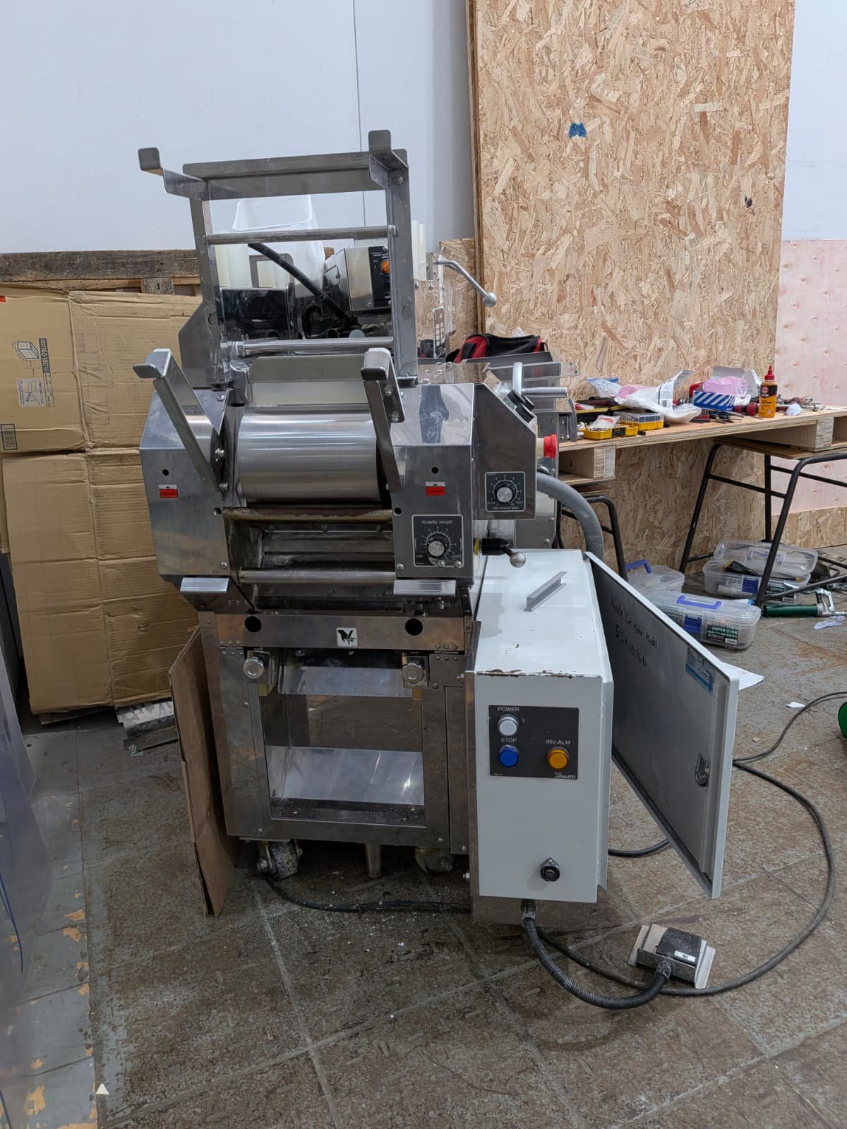 Stainless steel kitchen or industrial machine with control knobs, levers, and a transparent lid, situated on a cluttered workspace with tools, boxes, and cardboard in the background.