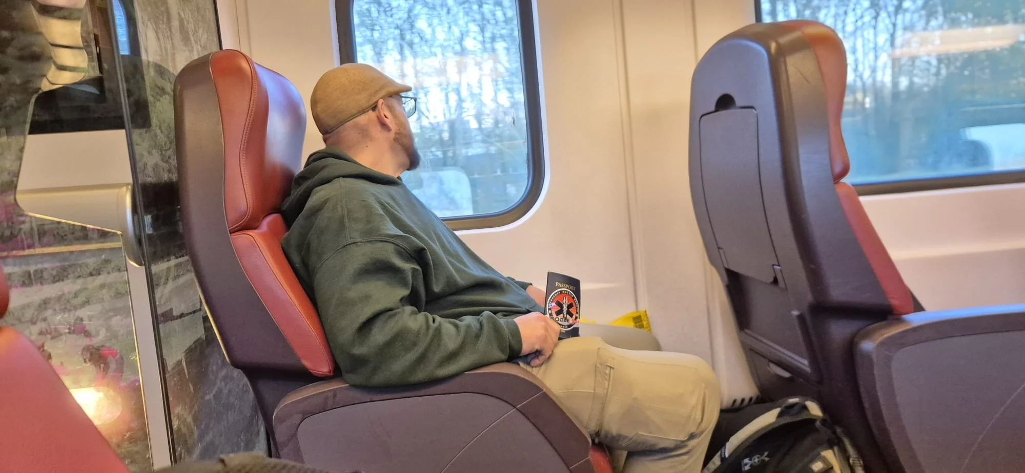 A man with a beard, wearing glasses, a brown cap, a green hoodie, and khaki pants, is sitting with his eyes closed on a train seat, holding a passport. He appears to be resting or sleeping.