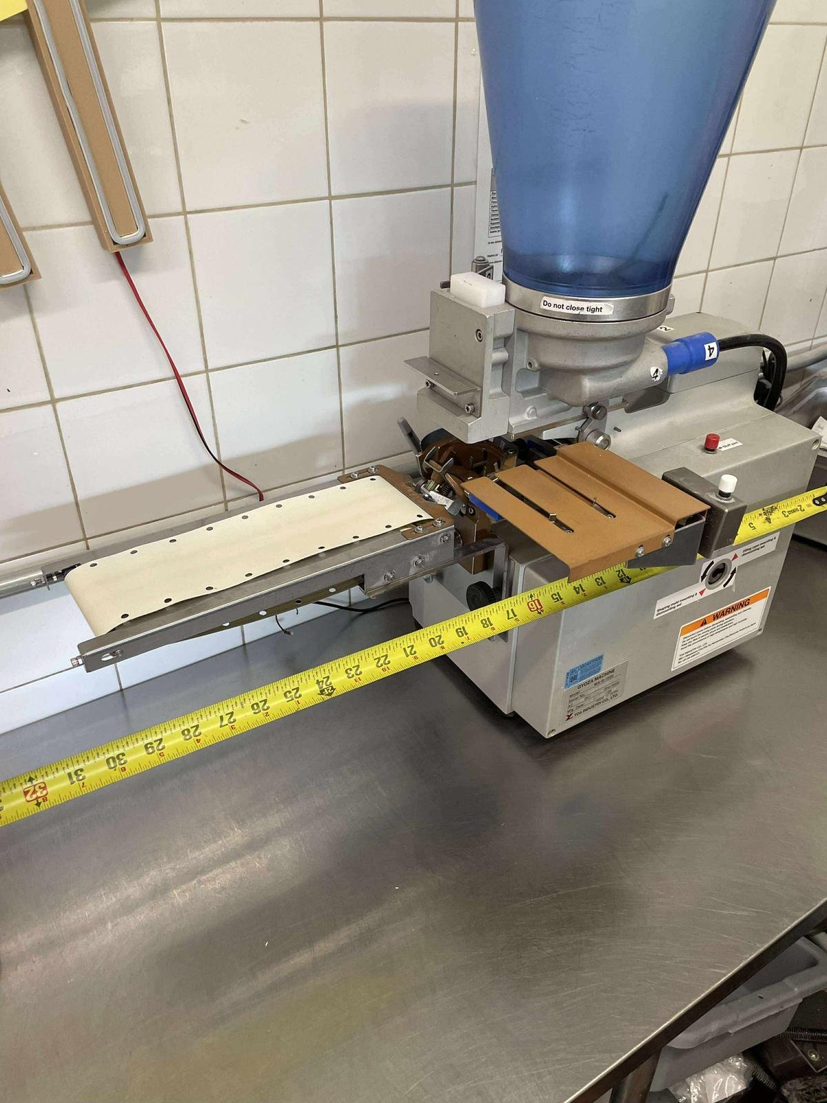 A pharmaceutical or laboratory tablet counting machine with a blue hopper, a conveyor belt, and measurement tape on a stainless steel table.
