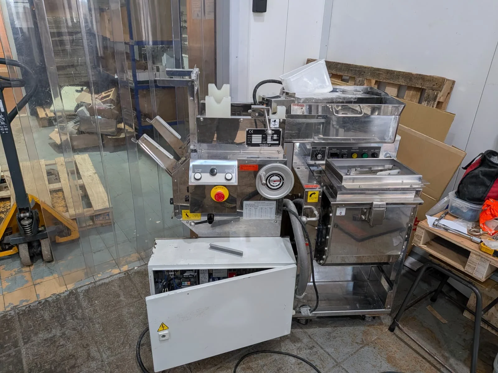 Industrial food processing machine made of stainless steel, with controls, dials, and a plastic bag holder, placed on wheels in a storage or kitchen area with cardboard boxes and other equipment nearby.
