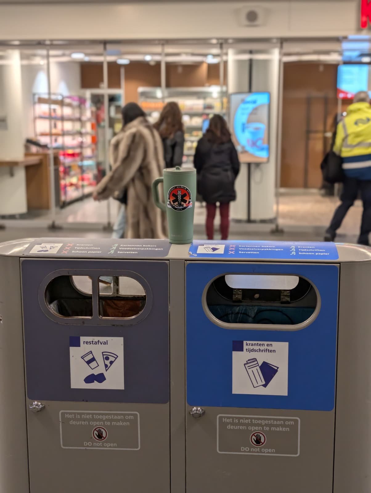 A public space with a gray trash bin with two compartments for trash and recycling labeled in Dutch, with a coffee mug with a medical symbol on top, in front of a shopping mall entrance where people are entering and exiting.