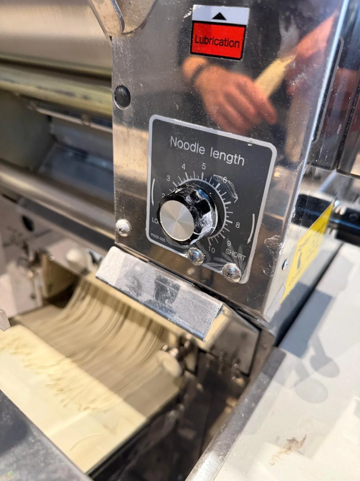 A pasta noodle cooker with a metal control panel labeled 'Noodle length' and a dial set to approximately 4.5. The panel has a red label marked 'Lubrication' and a small black triangle indicator. The noodles are pouring out of the machine.