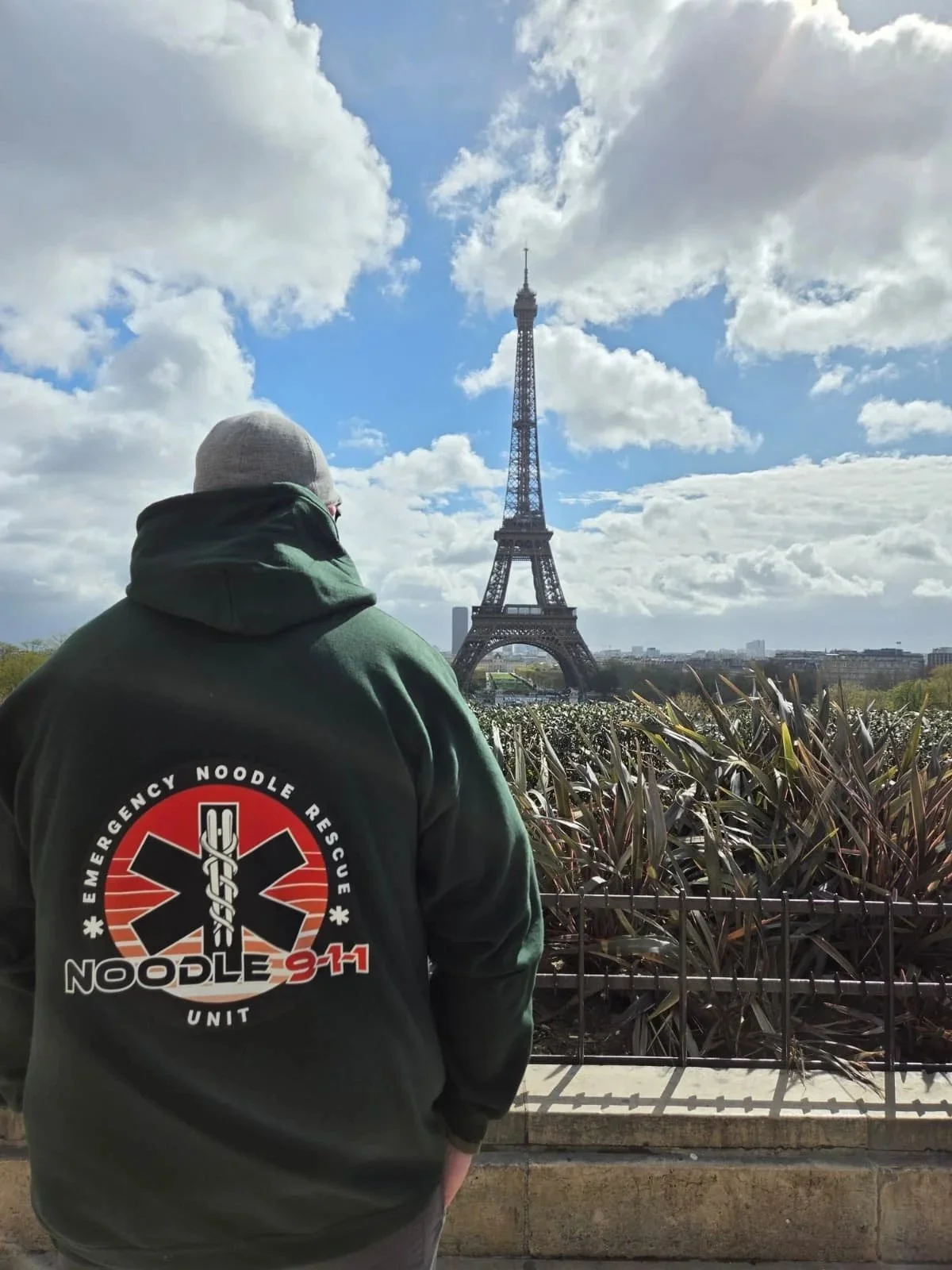 Person wearing a dark green hoodie with emergency noodle rescue patch on the back, standing and looking at the Eiffel Tower in Paris on a partly cloudy day.