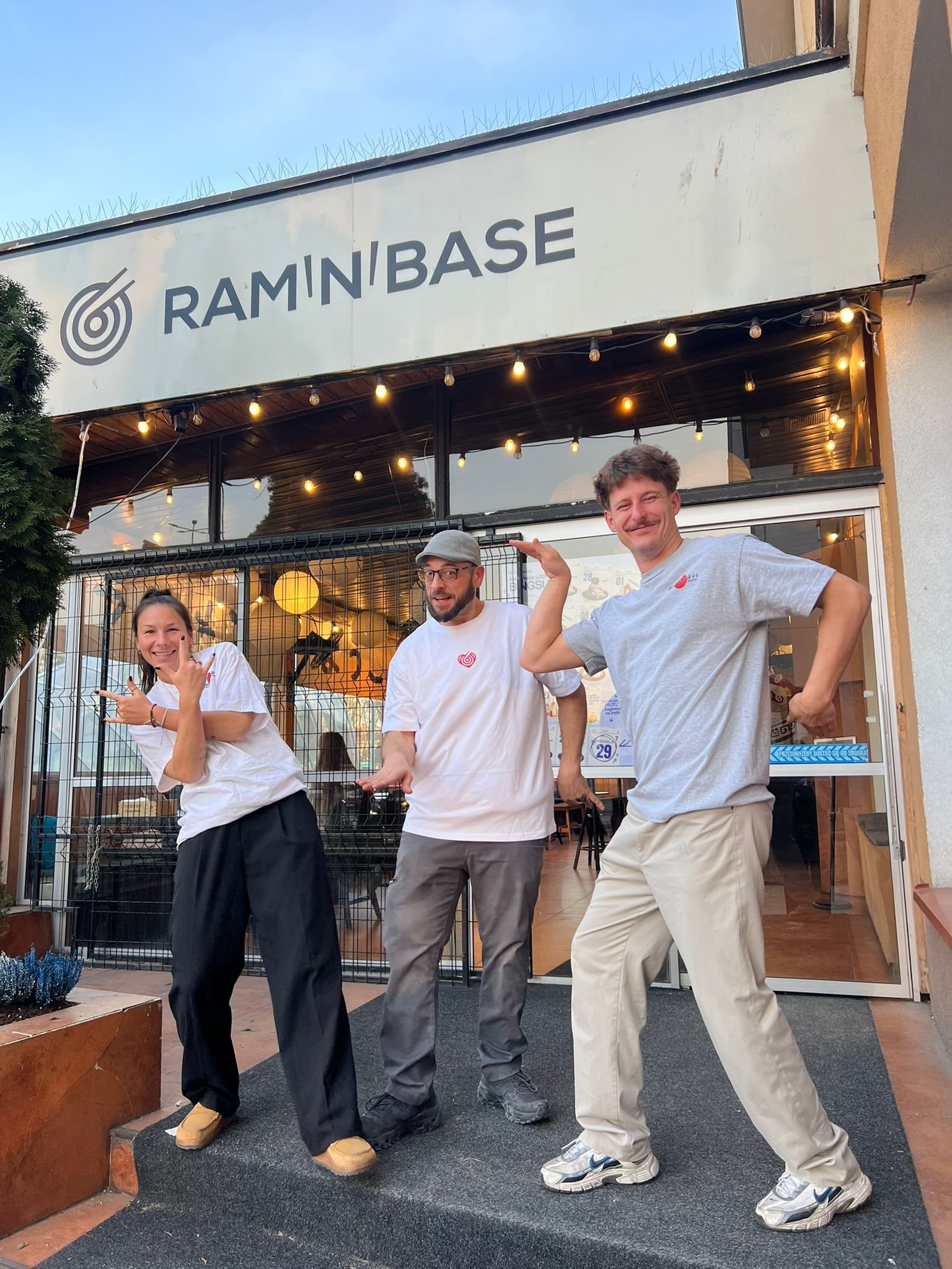 Three people standing outside a building with a sign that says 'Ramin'Base' and smiling, with two of them flexing their muscles and the third pointing at them.