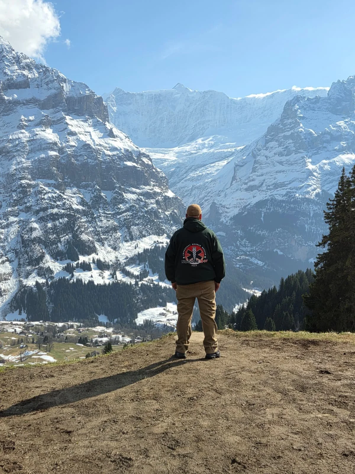 A man standing on a dirt path, overlooking snow-capped mountains and a small village below, with a clear blue sky overhead.