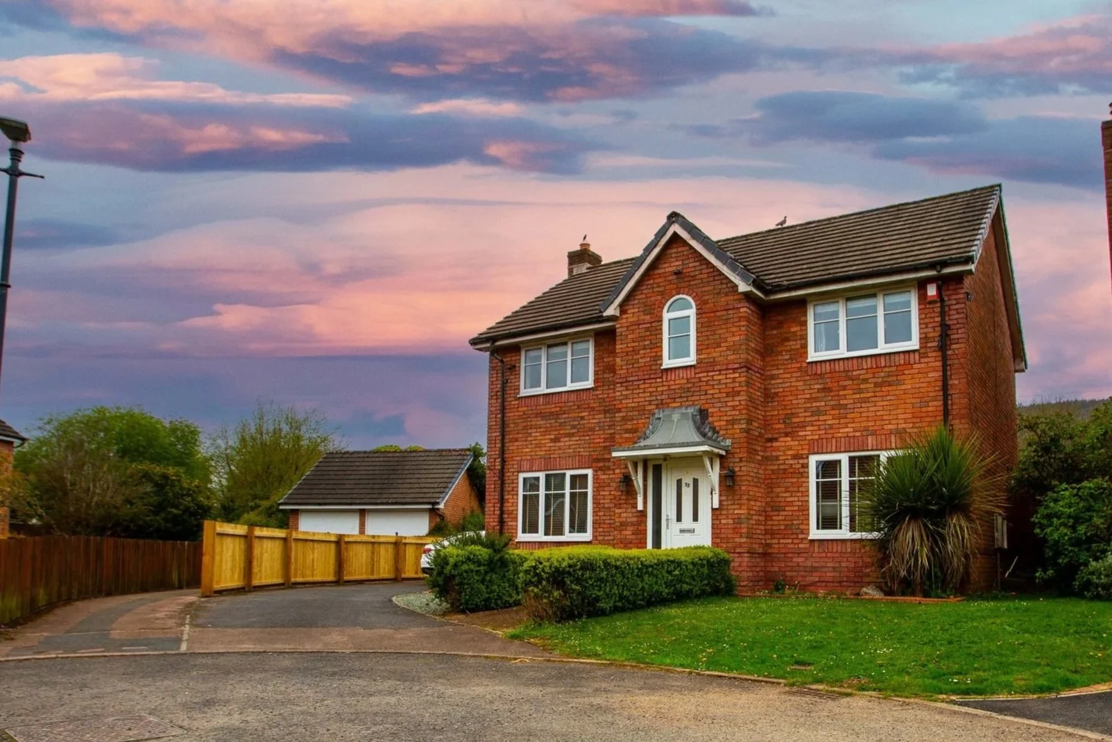 A quiet suburban street with brick houses, a small front yard with green plants, and a large sky with pink and purple clouds at sunset.