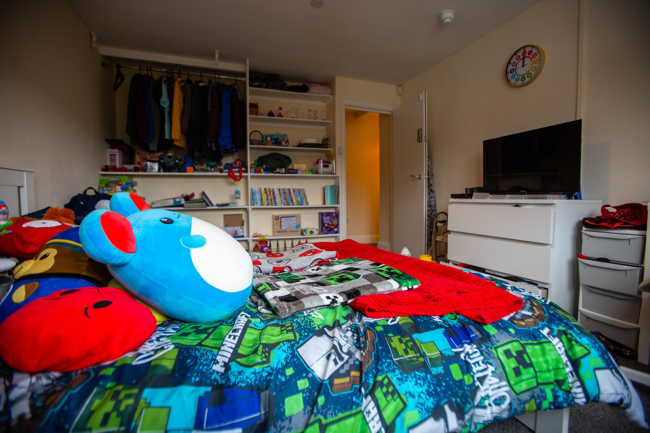 Child's bedroom with bed covered in colorful gaming-themed blanket, stuffed animal, and toys. Wall shelves with books and items, closet with hanging clothes, dresser, and a TV.