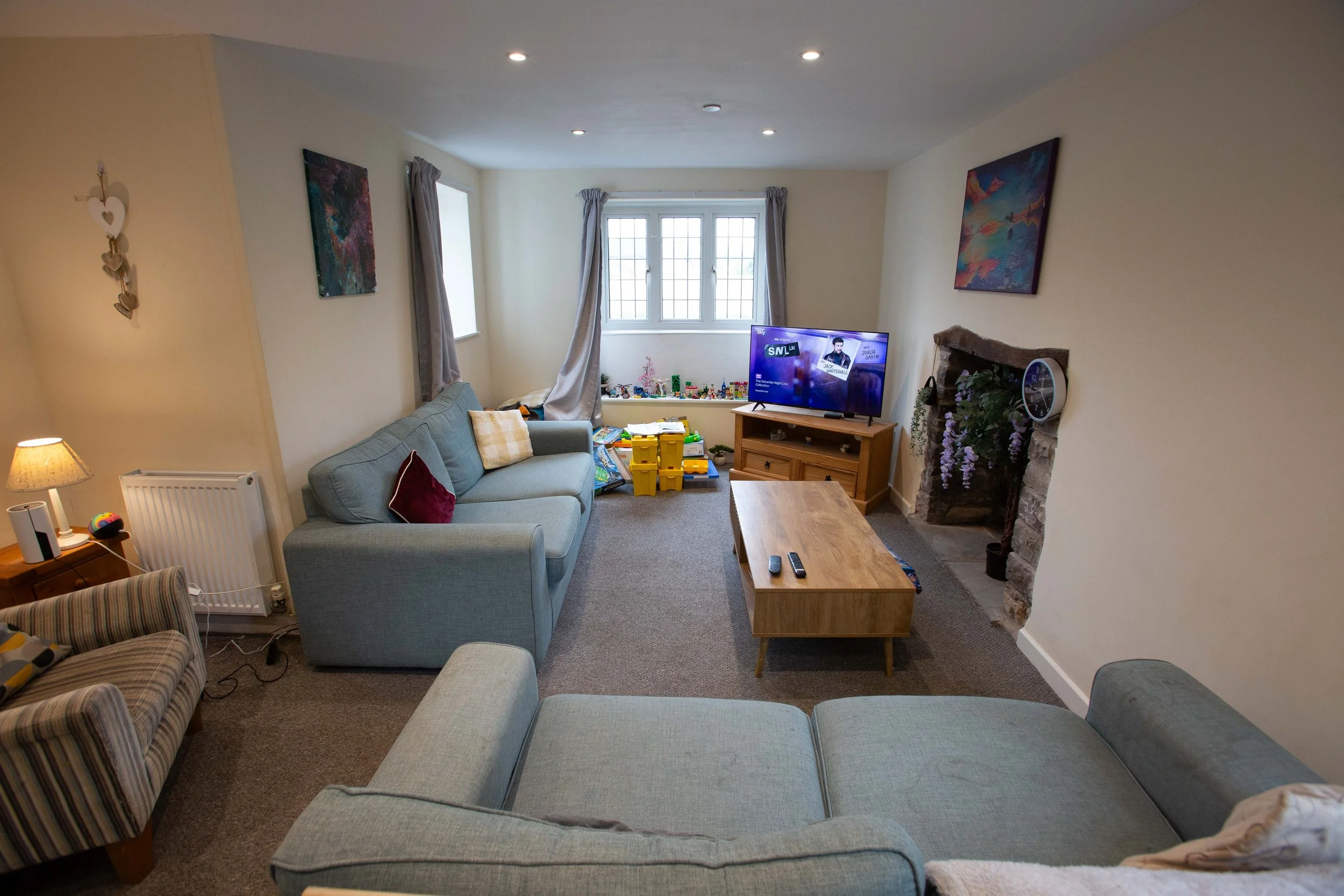 Living room with a light blue sectional sofa, a wooden coffee table, a flat-screen TV on a wooden stand, a stone fireplace with purple flowers, framed artwork on the walls, and toys near the window.