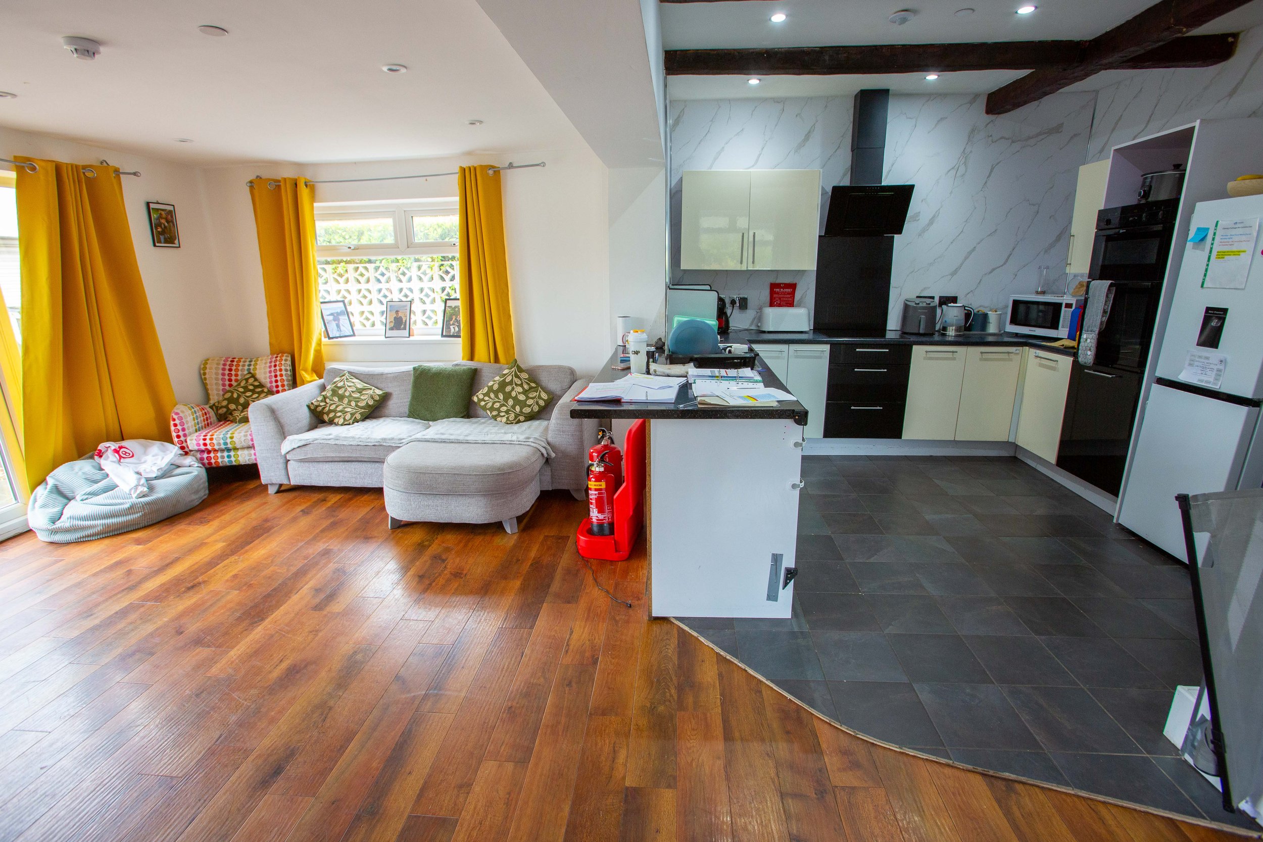 Open-plan living room and kitchen with yellow curtains, a grey sofa with multiple cushions, a window with framed photos, a kitchen counter with various appliances, and a fire extinguisher near the divider. The flooring is wood, and the kitchen has black and white cabinetry.