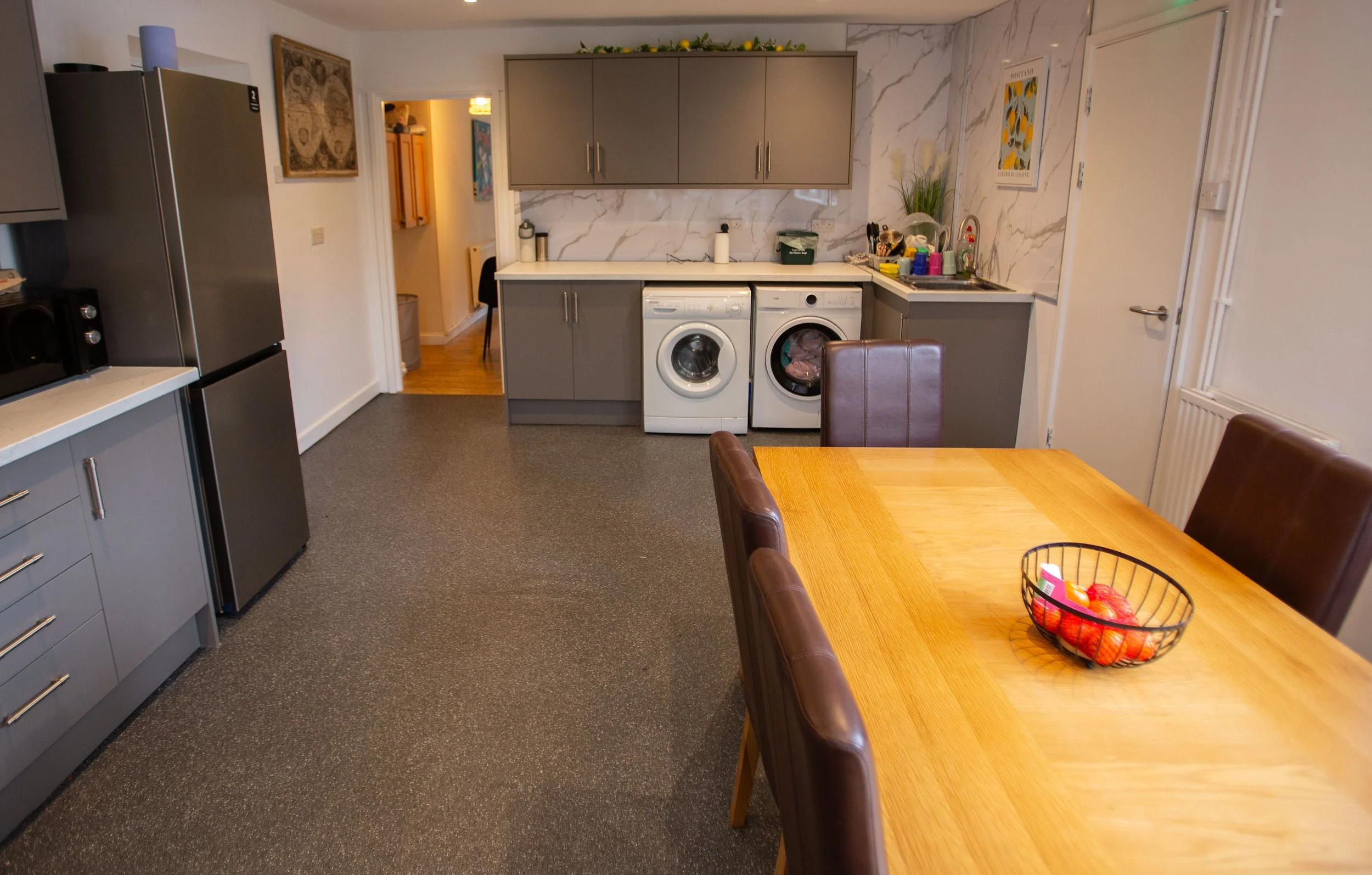 A kitchen with gray cabinets, a white marble backsplash, a refrigerator, a microwave, and a washing machine and dryer. There is a wooden dining table with brown chairs, a fruit basket, and various kitchen items near the sink.