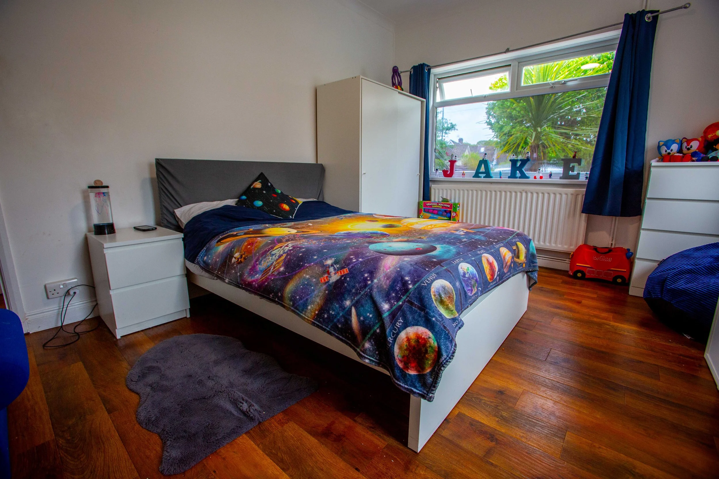 Child's bedroom with space-themed bedspread, white nightstand with lamp and phone, white wardrobe, window with blue curtains, toys on dresser, and a gray rug on wooden floor.