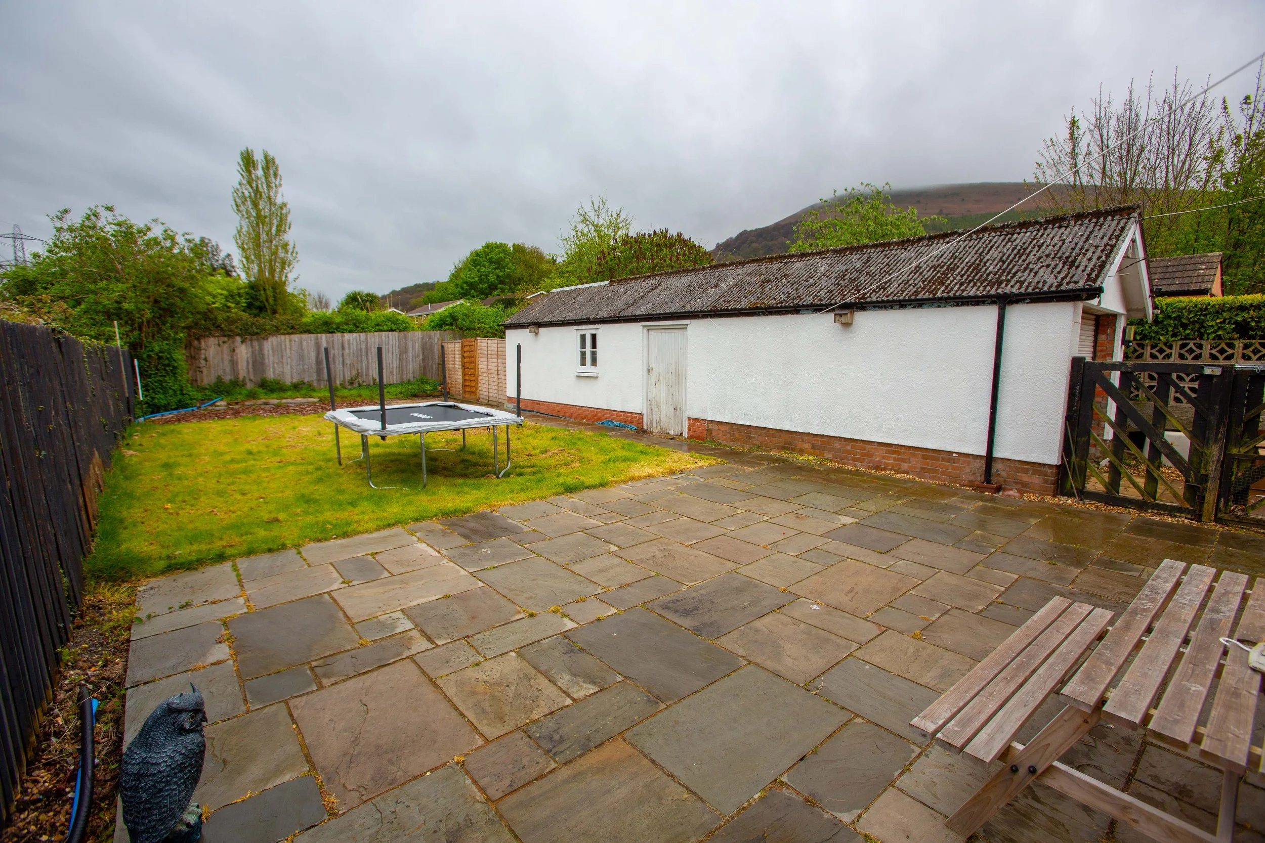 A backyard patio with large stone tiles, a white shed with a black roof, a trampoline, a wooden bench, and a wooden fence. Green trees and hills are in the background under a cloudy sky.