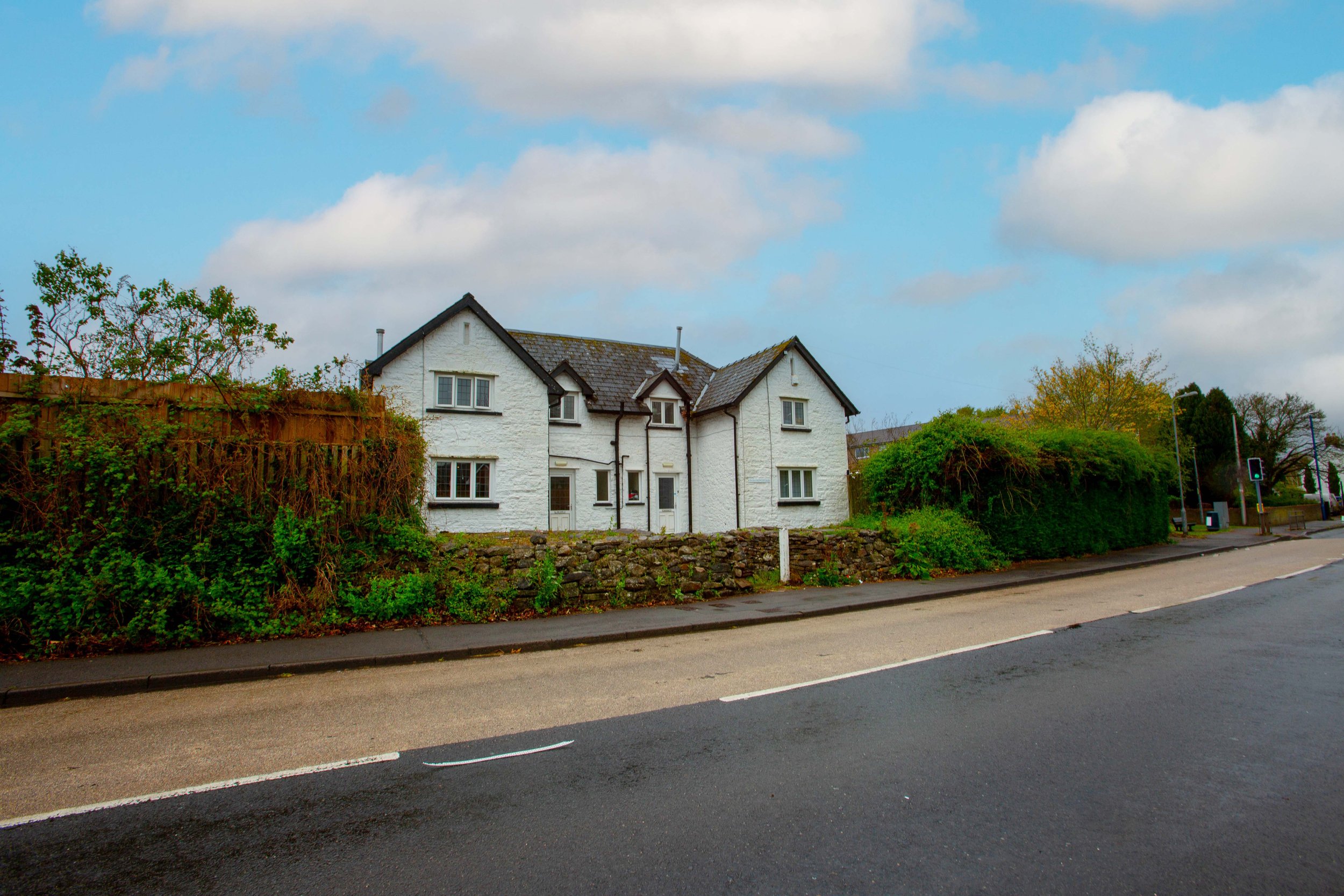 Residential street scene with white house, green trees and bushes, cloudy blue sky, and a traffic light.