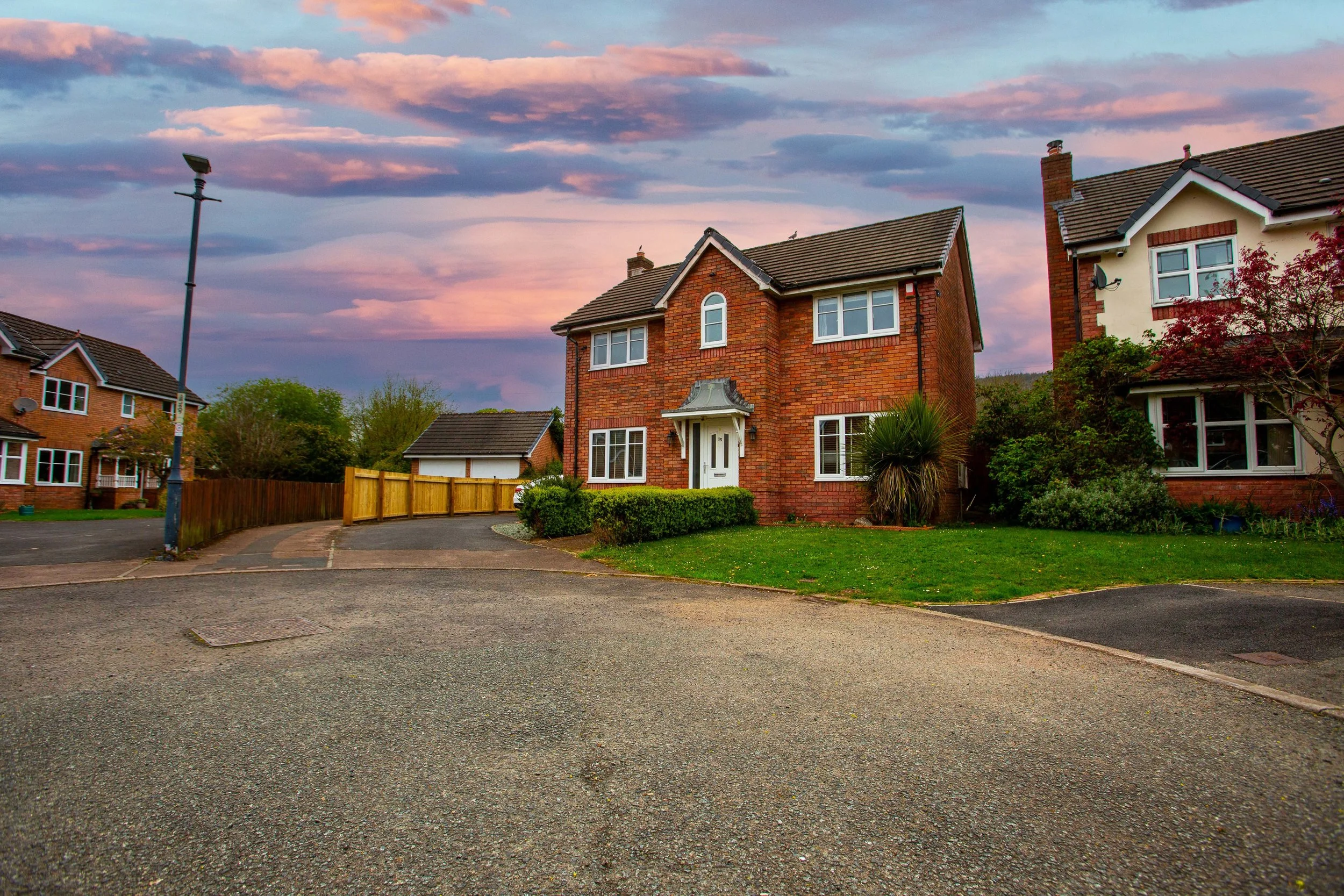 A quiet suburban street with brick houses, a small front yard with green plants, and a large sky with pink and purple clouds at sunset.