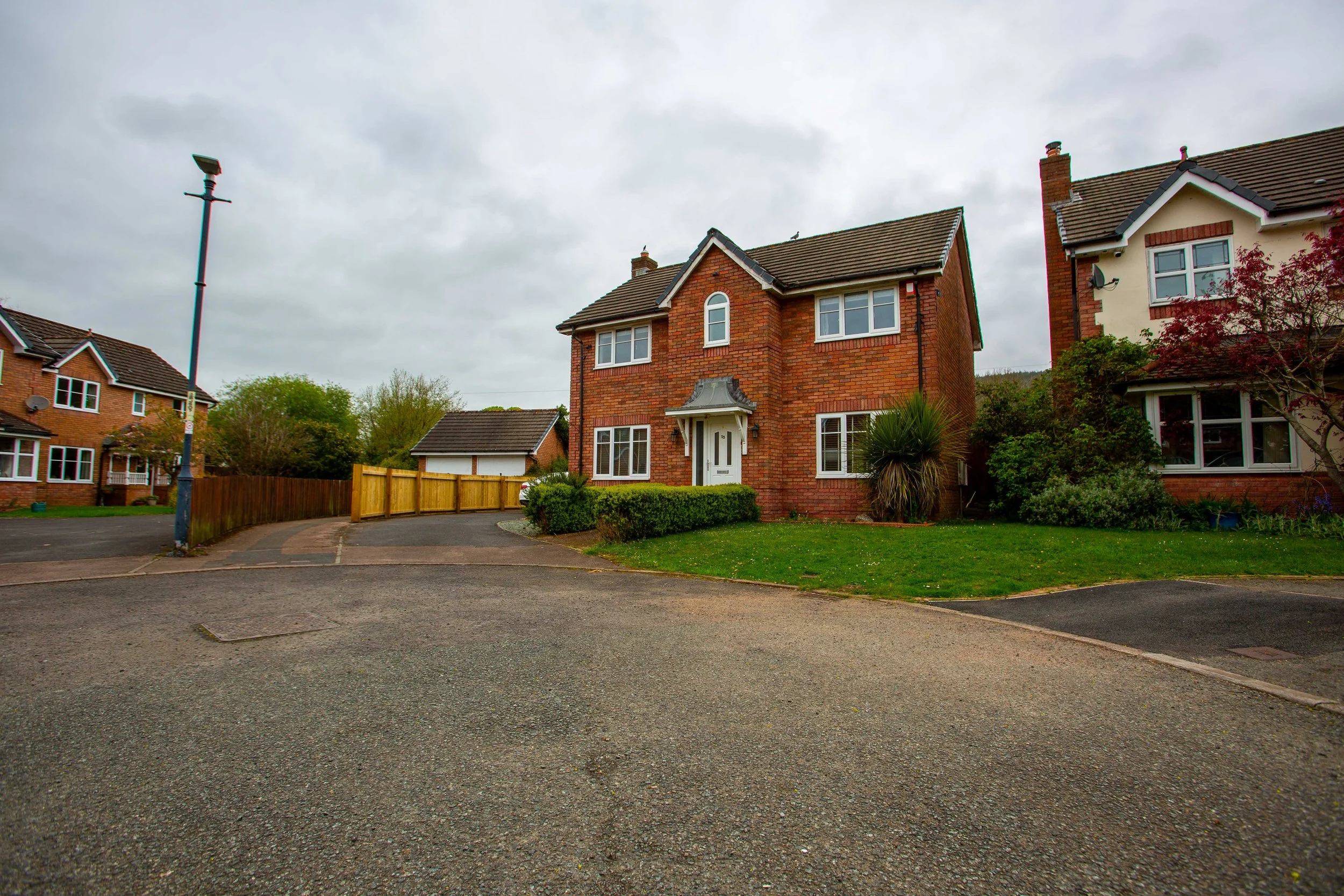 View of a residential neighborhood with brick houses, a driveway, a grass lawn, trees, and a cloudy sky.
