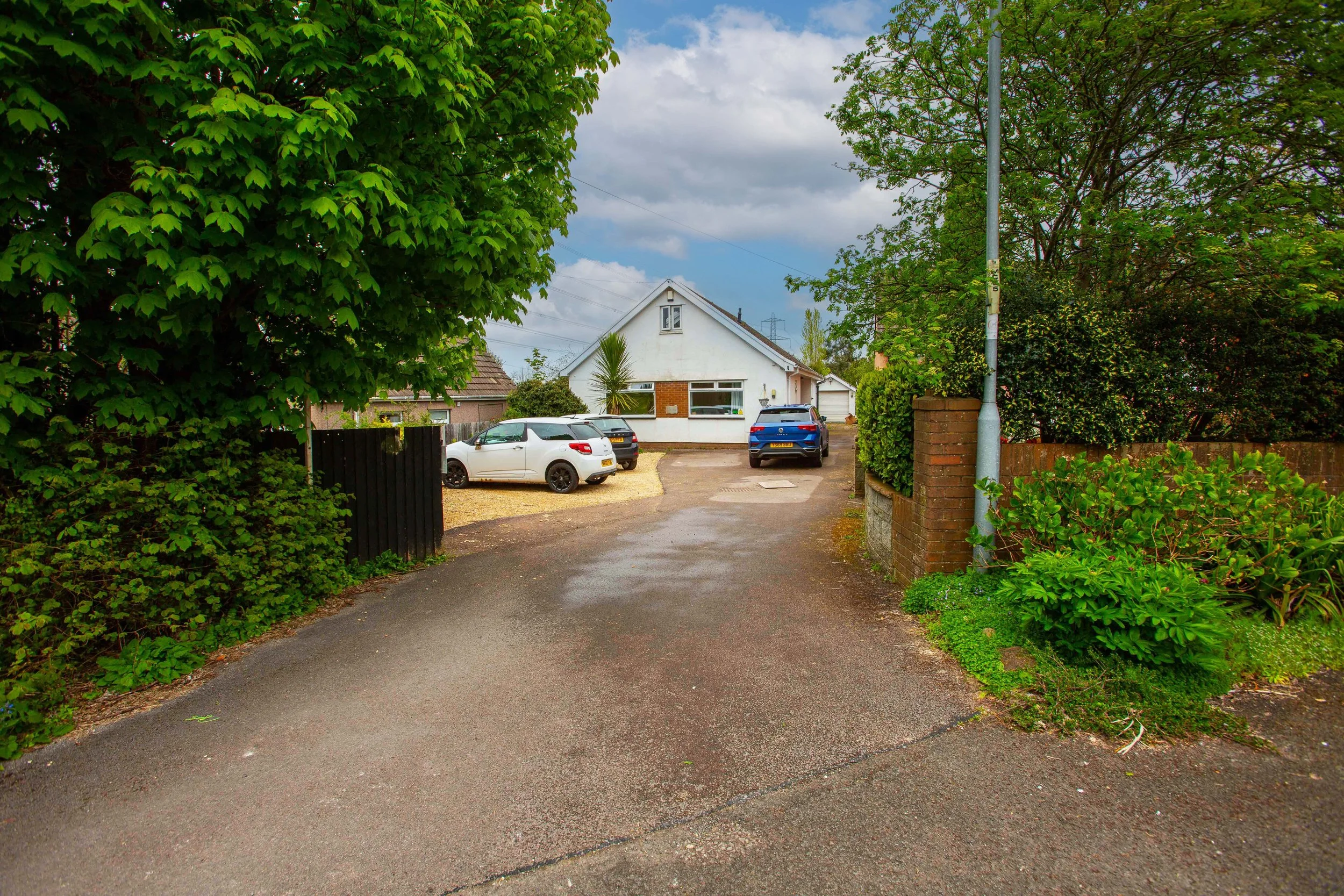 View of a suburban house with a driveway and parked cars, surrounded by lush green trees and bushes, under a partly cloudy sky.