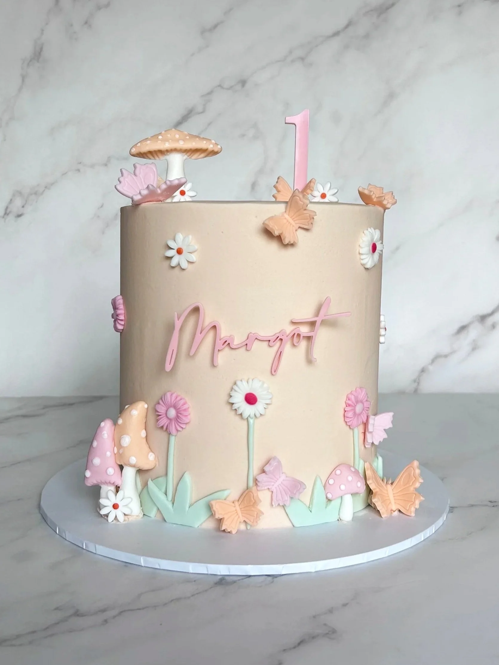A pastel-colored birthday cake decorated with fondant flowers, mushrooms, and butterflies, featuring a pink number one candle.