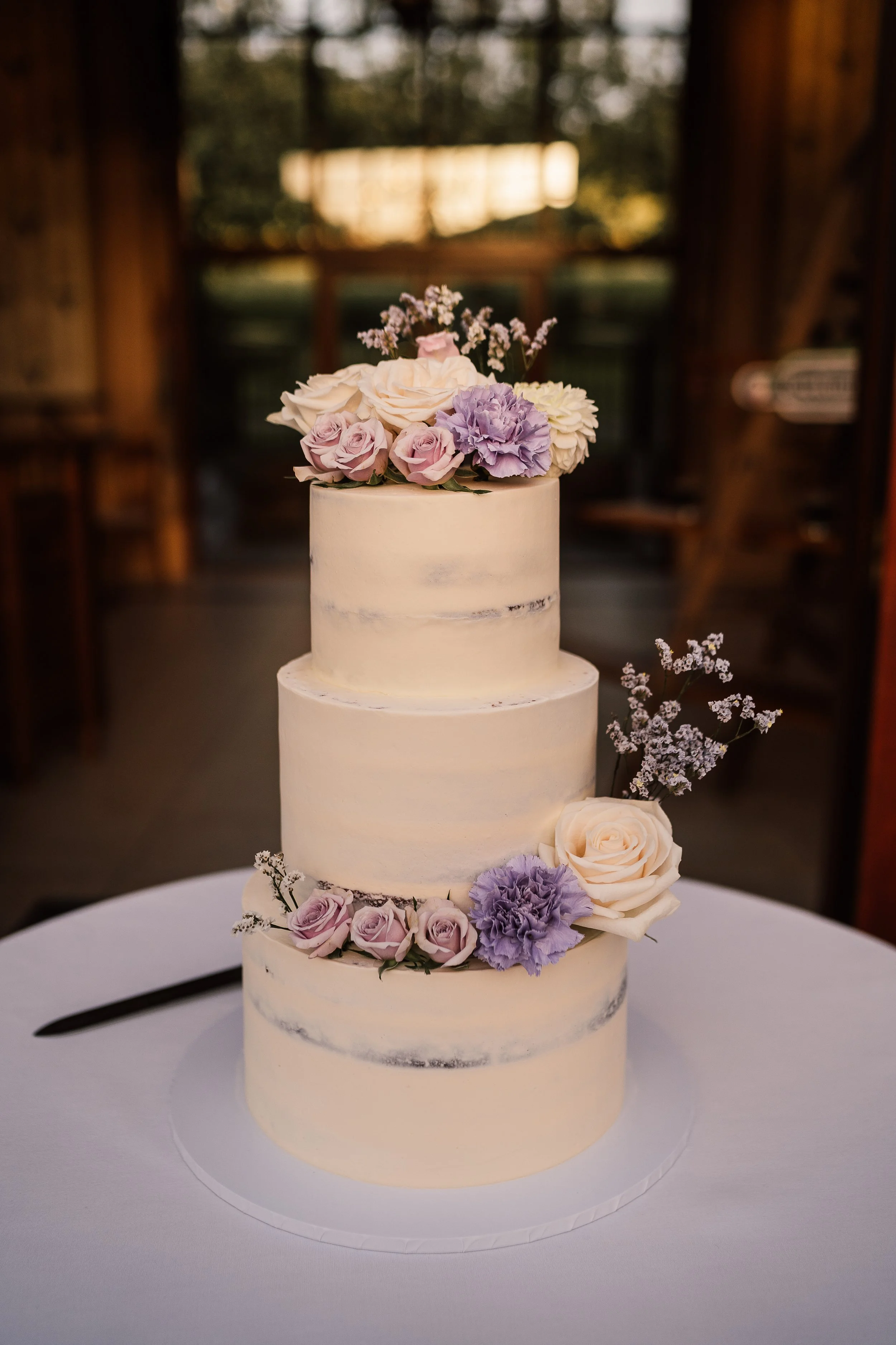 A three-tiered wedding cake decorated with light purple, white, and pastel pink roses, lavender, and small white flowers, set on a white table with a brown wooden background.