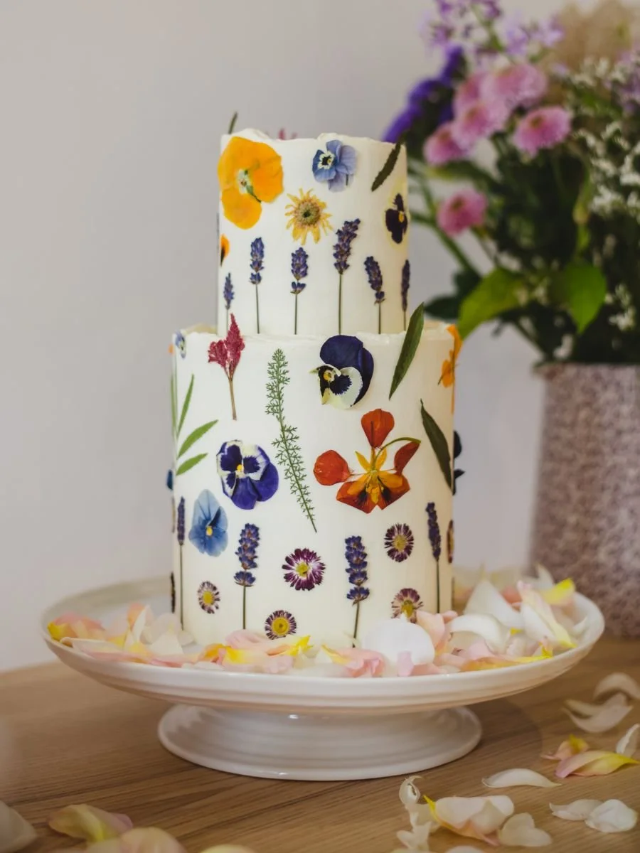 Two-tiered white cake decorated with pressed flowers including pansies, lavender, and various other colorful flowers, on a white cake stand surrounded by scattered flower petals, with a large bouquet of pink and purple flowers in the background.