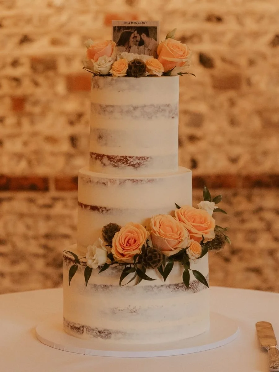 A three-tier semi-naked wedding cake decorated with pink roses, white flowers, pinecones, and greenery, topped with a photo of a couple and a sign reading 'Mr & Mrs Grant'.