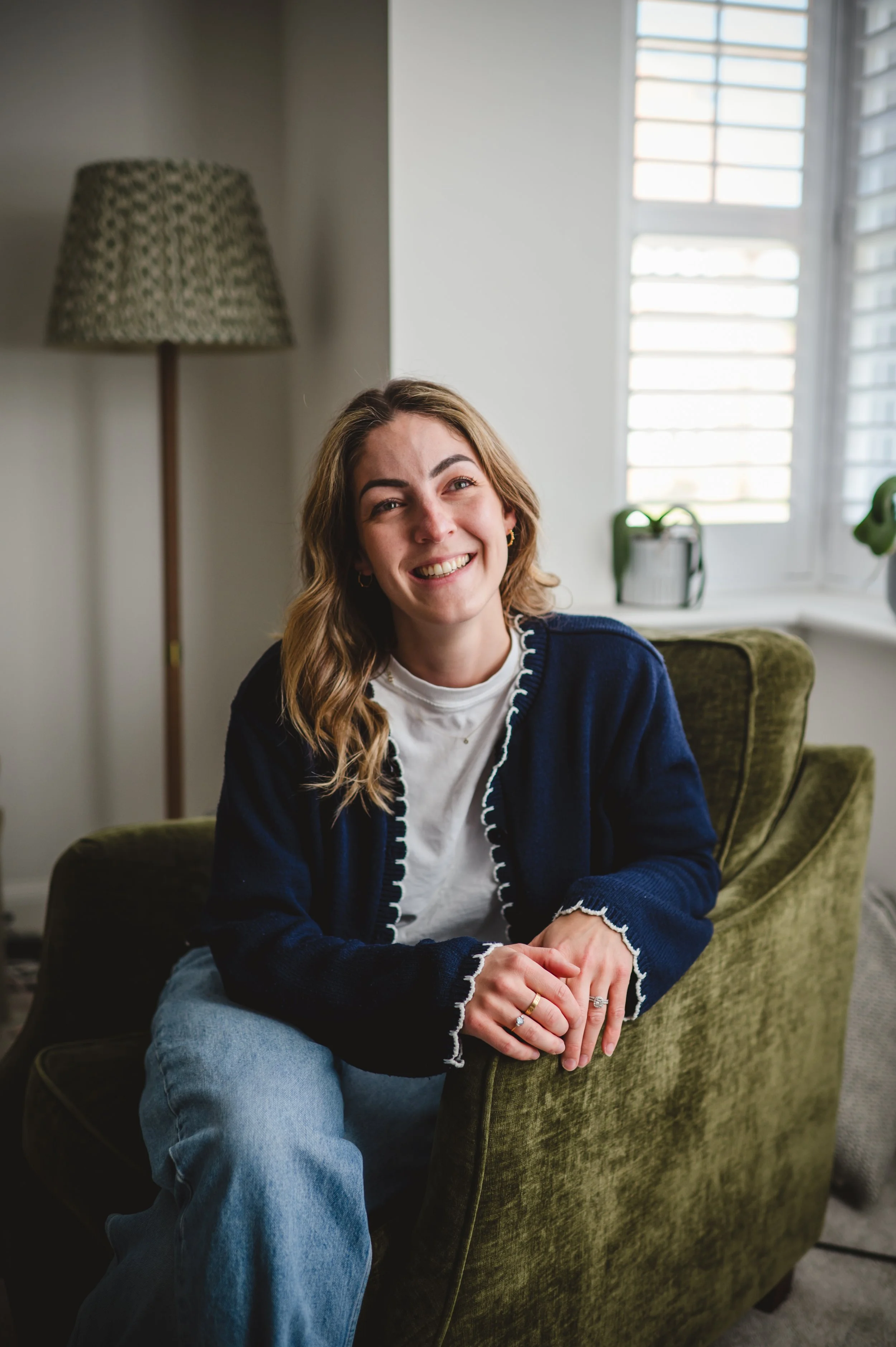 A young woman with wavy, shoulder-length hair smiling and sitting on a green velvet armchair in a bright room with white walls, a window with blinds, and a tall floor lamp with a patterned shade.