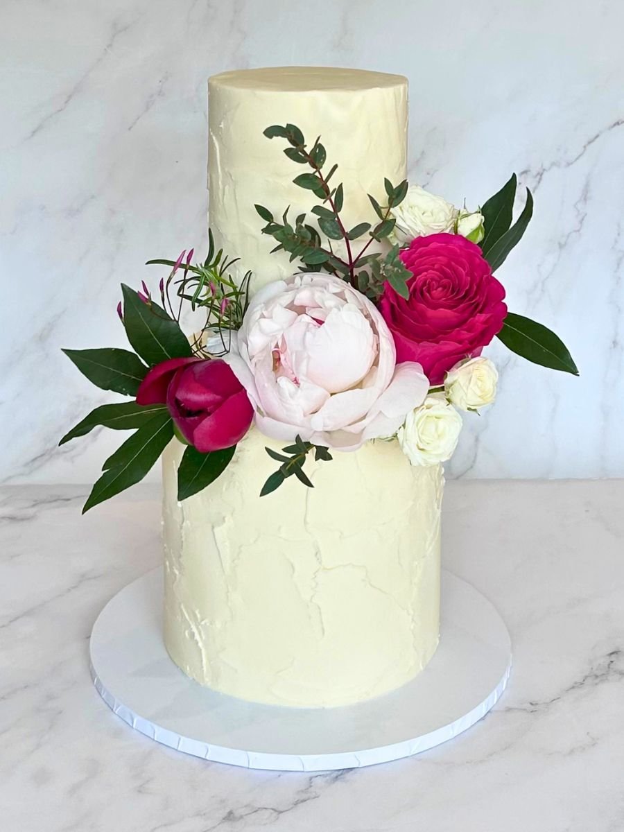 Two-tiered cake with white frosting topped with pink and white peony flowers and dark green leaves, set on a white cake board.