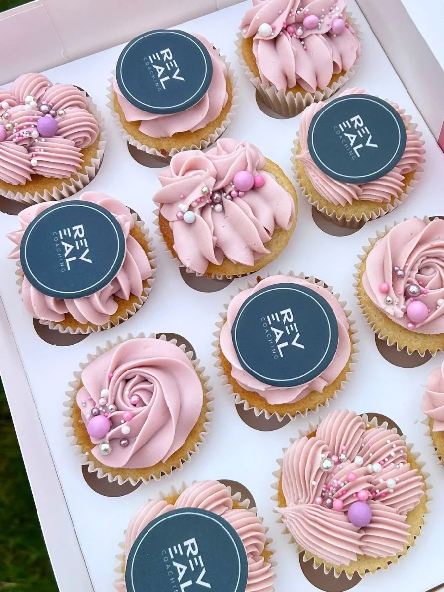 A box of cupcakes decorated with pink frosting, pearl and spherical sprinkles, some with printed round toppers that say 'REV REAL' in white text on a dark background.