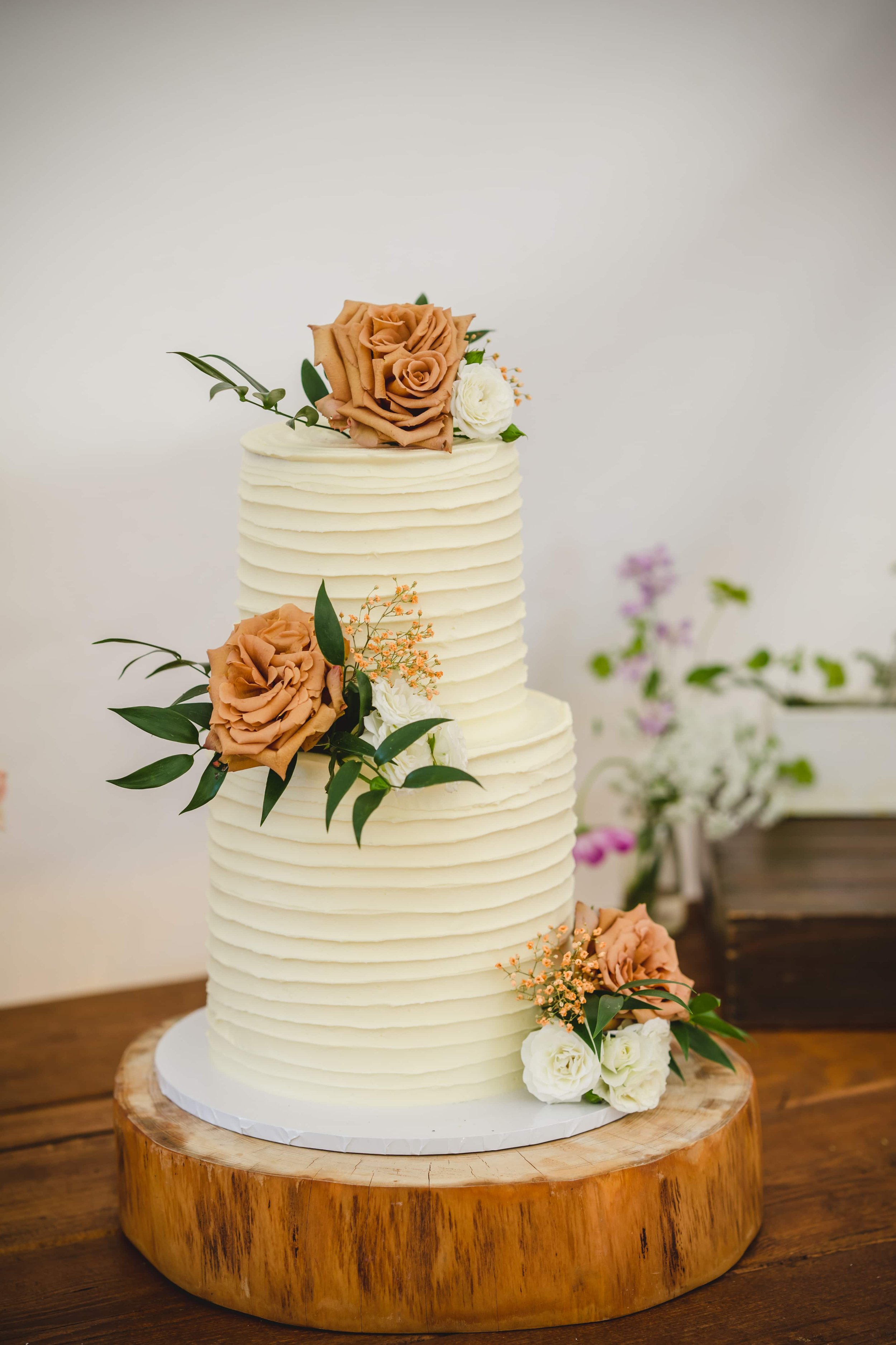 A white, textured, tiered wedding cake adorned with peach, white, and pink flowers, small leaves, and gold accents, placed on a wooden cake stand with a rustic background of purple and white flowers.