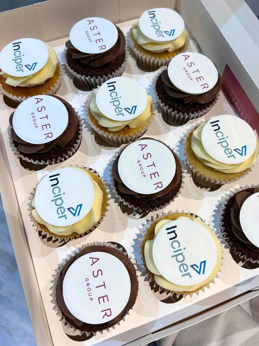 A box of colorful cupcakes topped with edible logos of the Inciper and Aster Group companies.
