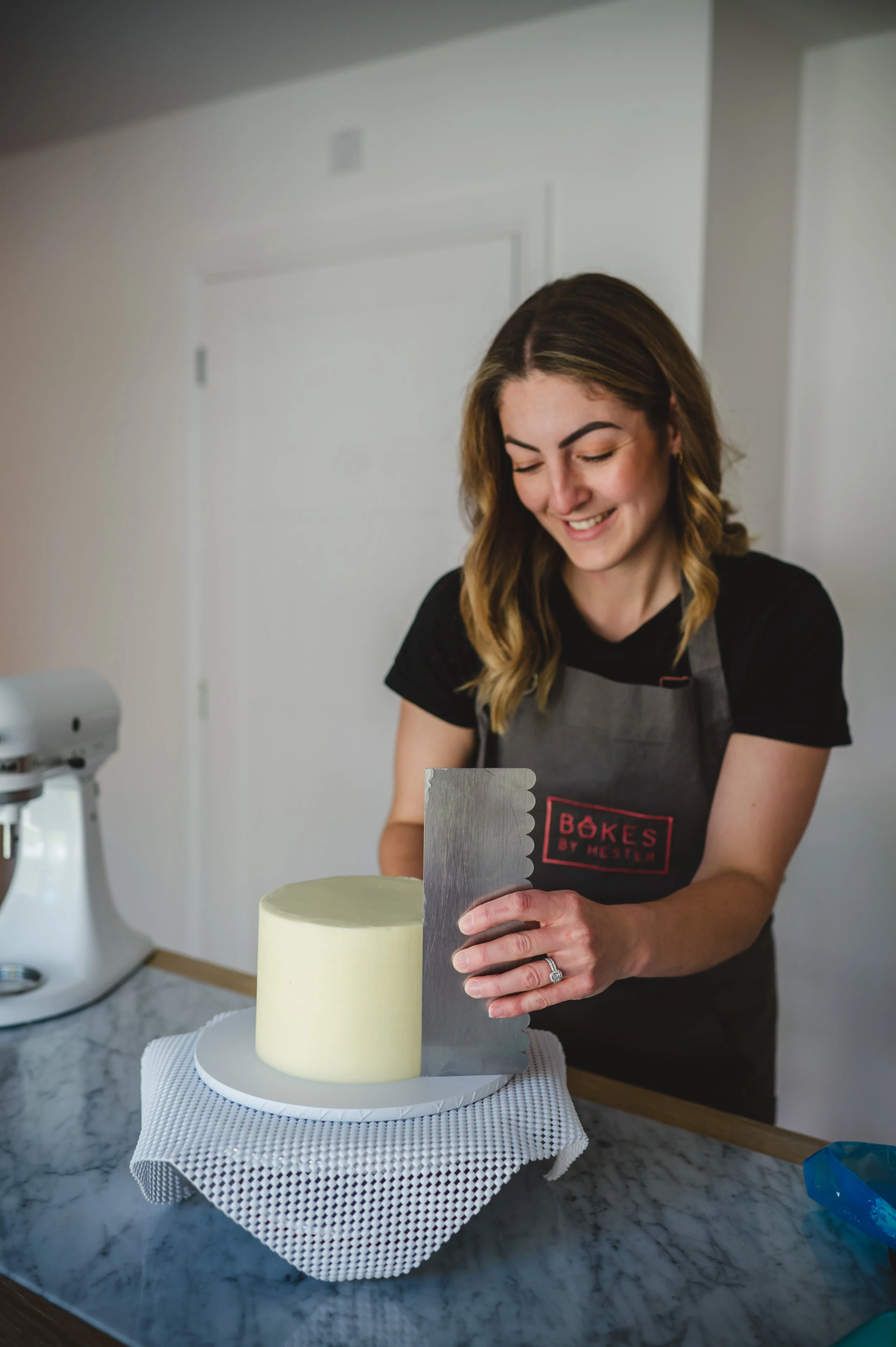 A woman wearing a black T-shirt and gray apron is smiling while using a cake spatula on a white cylindrical cake. The cake is on a white textured cake board, and the scene is set on a marble countertop in a kitchen.