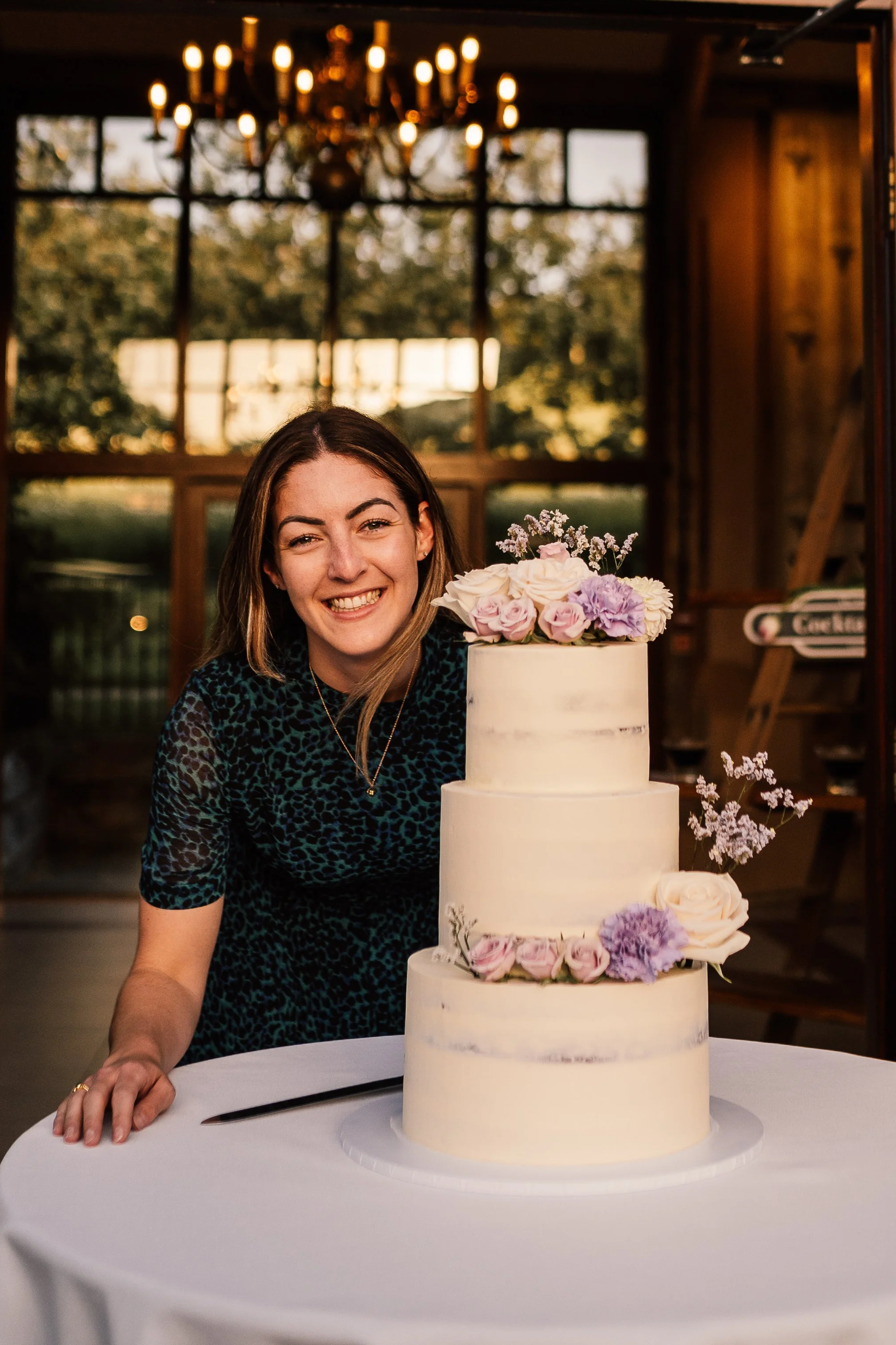 A smiling woman with shoulder-length brown hair wearing a blue leopard print top, standing next to a white tiered wedding cake decorated with pink and purple flowers, in a warmly lit indoor venue with large windows and a chandelier.