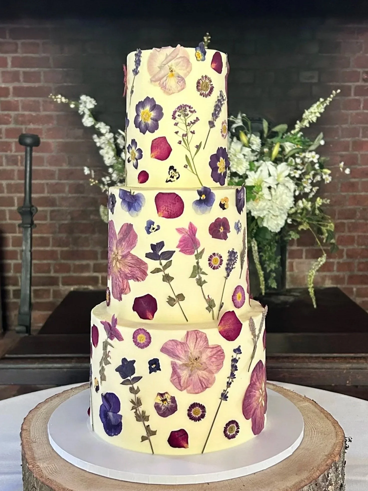 Three-tiered cake decorated with edible dried flower petals, displayed on a wooden slab with a white base, with white floral arrangements in the background against a brick wall.