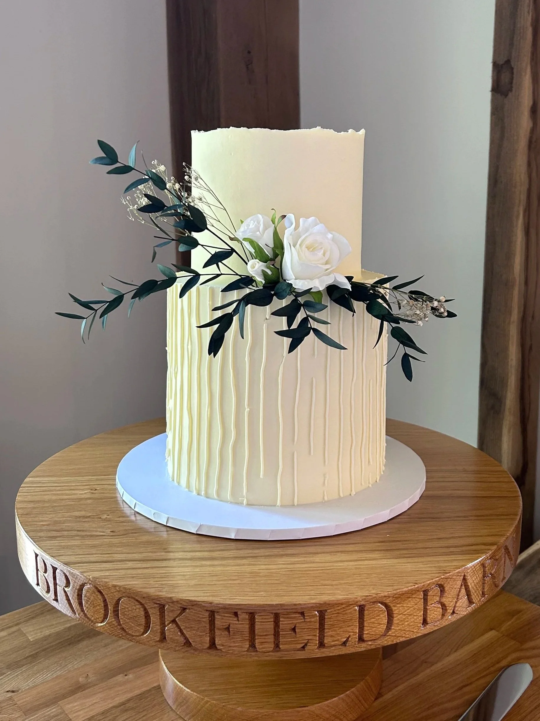 A tall white cake with a textured vertical stripe pattern, decorated with white roses and green foliage, on a round wooden cake stand with engraved lettering.