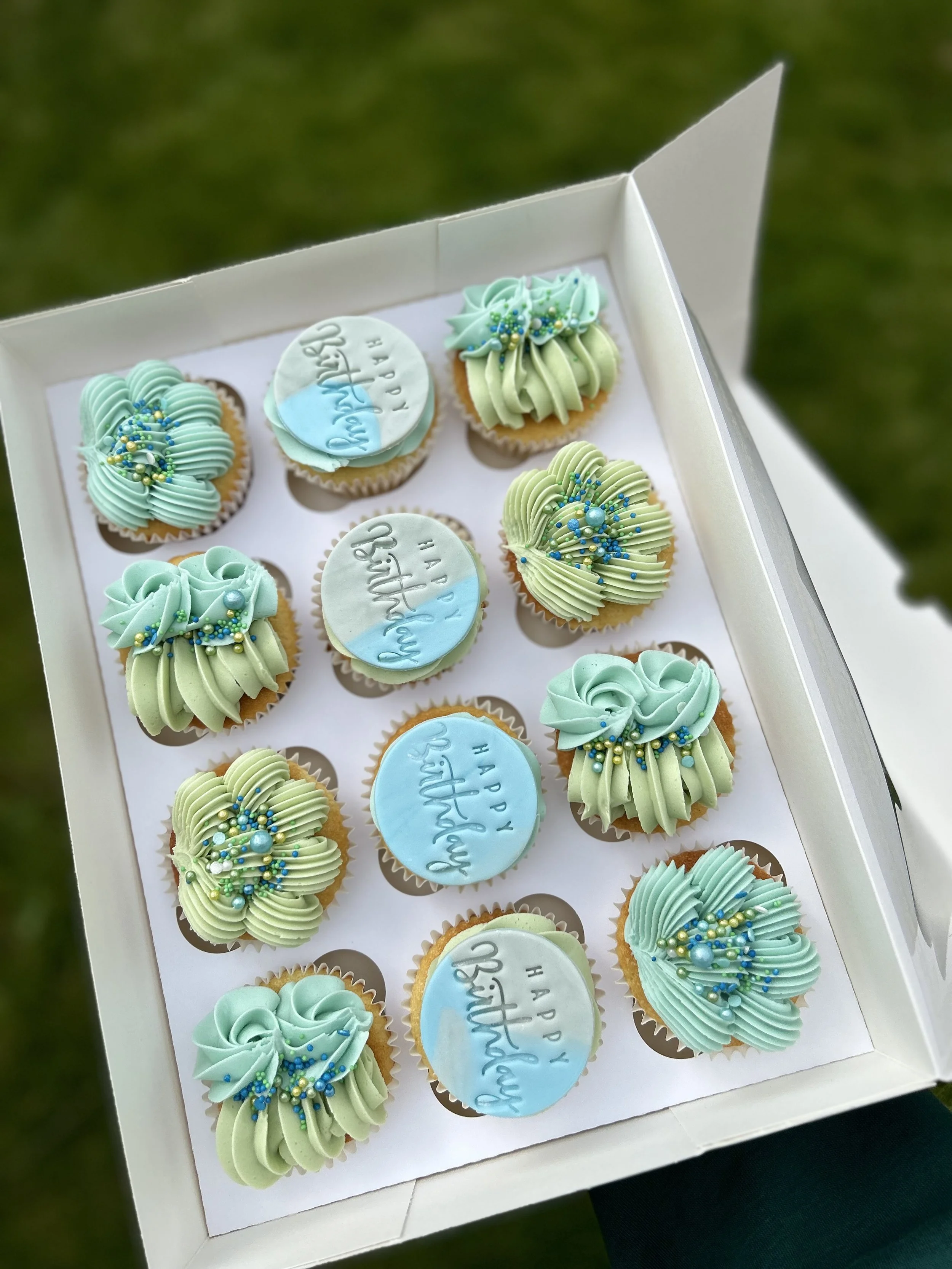 A box of decorated cupcakes with pastel green and blue frosting, sprinkles, and round toppers that say "Happy Birthday."