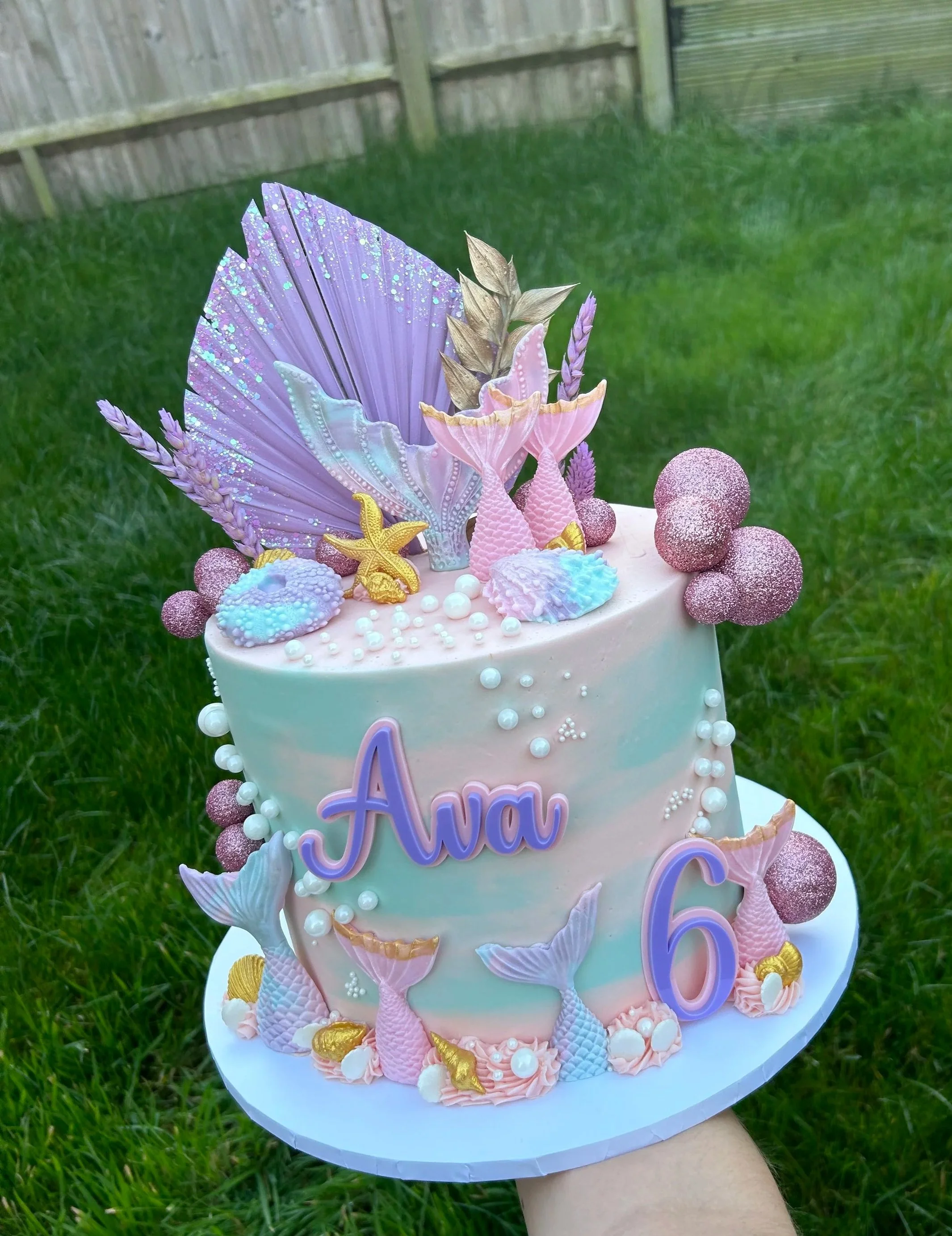 Decorative birthday cake with pastel colors, seashells, starfish, and ocean-themed decorations, with the text 'Aya 6' on it, held outdoors on grass.